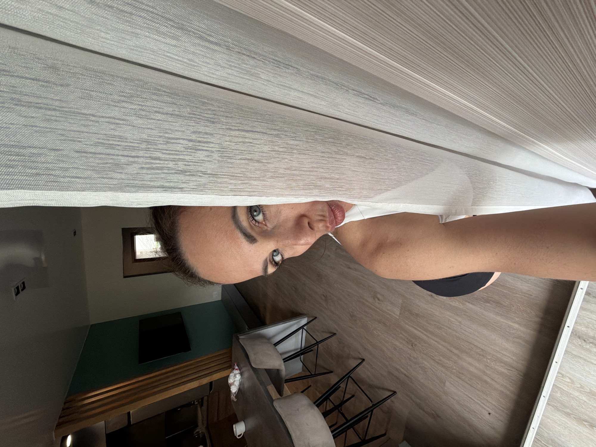 The image shows a woman peeking from behind a white sheer curtain. She has light skin, blue eyes, and dark brown hair pulled back. She is wearing a white top and her lips are pursed. In the background, there is a dining area with a dark gray table and chairs, a wooden partition, and a television on a teal wall. The flooring is light wood. The scene appears to be an indoor residential setting, but its specific location in Birobidzhan, Russia, cannot be determined from the visual information provided.
