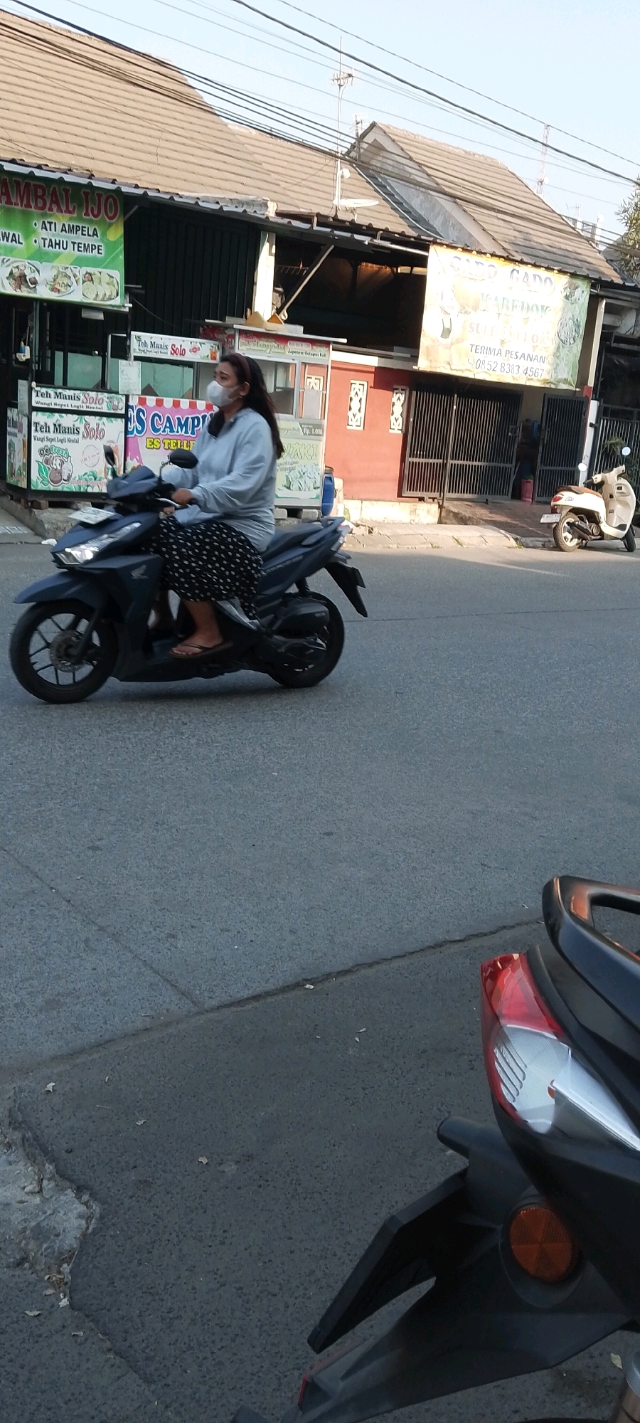 A woman wearing a face mask and a light blue hoodie with a black and white polka dot skirt rides a dark blue scooter down a paved road. She is wearing flip-flops. The scooter is in motion. In the background, there are storefronts with signage in Indonesian. One sign reads "AMBAL ILJO" and another reads "GADO GADO". A white scooter is parked in front of a building. The environment appears to be a street in Satriamekar, Indonesia. The sky is clear and the lighting suggests it is daytime.