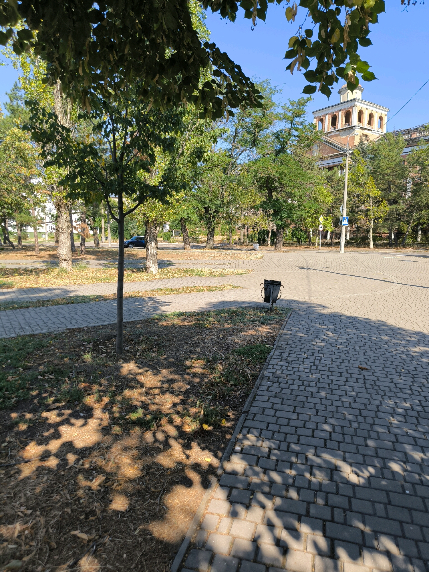 The image depicts an outdoor scene on a sunny day, likely in an urban park or public space in Mykolaiv, Ukraine. In the foreground, a paved walkway made of grey interlocking bricks curves into the distance, casting dappled shadows from overhead foliage. To the left of the walkway, a patch of dry grass and fallen leaves is visible, with a young tree growing from it. A black trash can with a decorative base stands near the edge of the walkway.
Further into the scene, another paved area extends outwards, forming a junction or plaza. A dark blue car is parked on the left side of this area. Beyond the paved areas, a park-like setting unfolds with numerous trees featuring green and yellowing leaves, suggesting it might be late summer or early autumn. In the background, a large, multi-story building with a distinct architectural tower featuring arched windows and a domed top is visible through the trees. Traffic signs and a lamppost are also present on the right side of the paved junction. The sky is clear and blue. The overall impression is one of a well-maintained public area under bright sunlight.