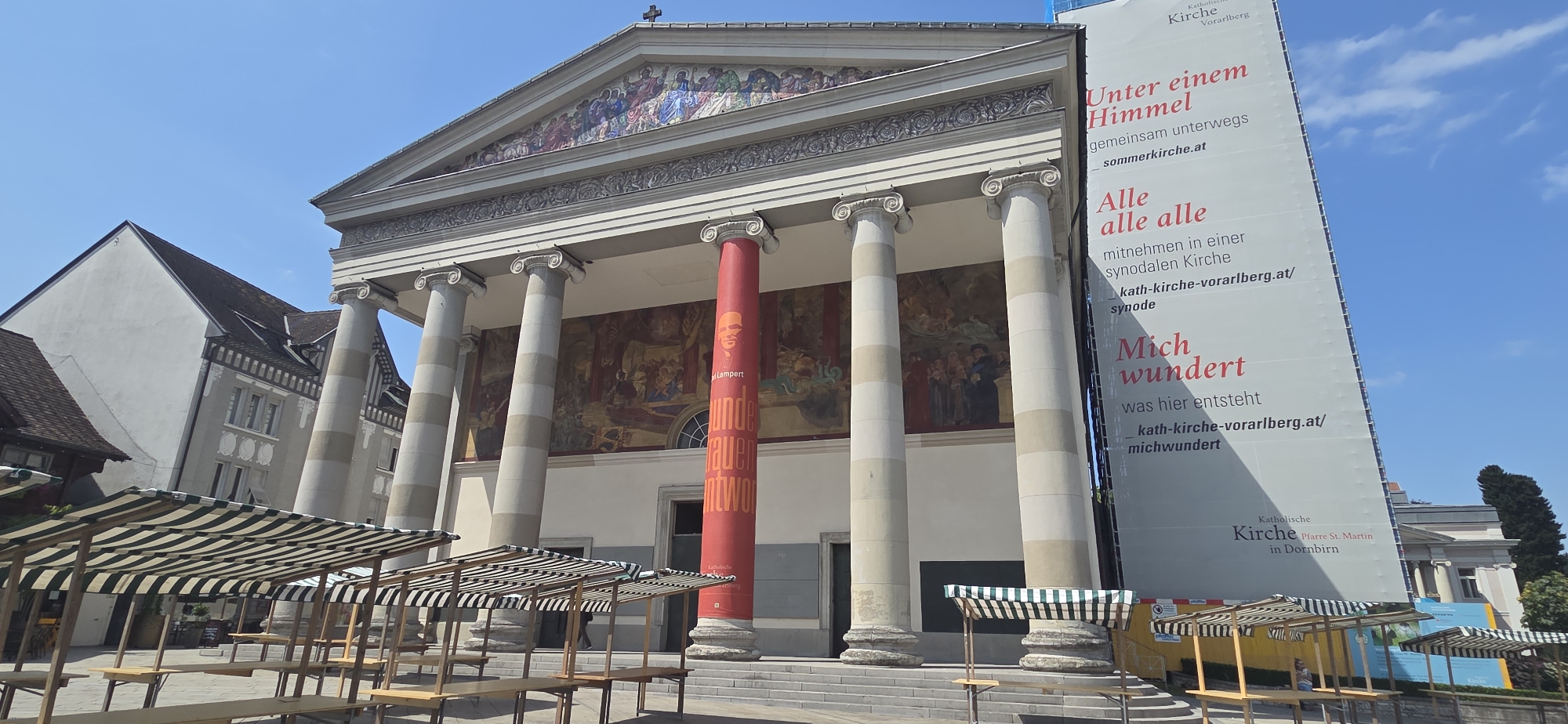 The image shows the Catholic Church of St. Martin in Dornbirn, Austria, on a clear, sunny day. The church features a grand Neoclassical facade with a pediment adorned with a mosaic and supported by large Ionic columns. A large red banner hangs from one of the columns, featuring an orange portrait and text. To the right of the church, a large white banner displays text in German related to the church's synod. In the foreground, several empty market stalls with green and white striped awnings are set up, suggesting an outdoor market is either in progress or being prepared. The sky is a bright blue with a few wisps of clouds.