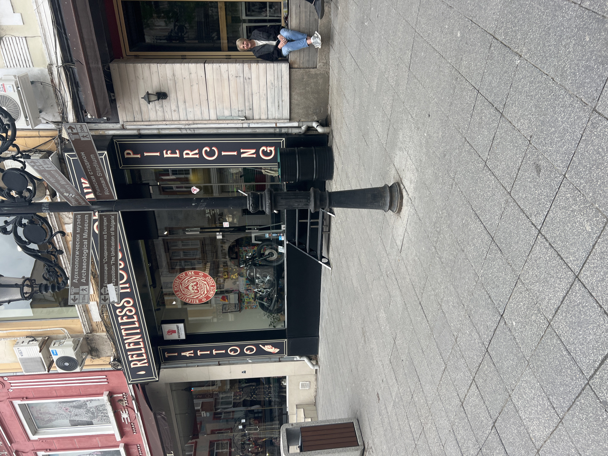 An outdoor street scene is captured during the daytime, likely overcast, with no direct sunlight. The image is vertically oriented but depicts a horizontal street view.
In the middle ground, a shopfront features prominent vertical signs displaying "PIERCING" and "RELENTLESS HOUSE TATTOO". A circular logo inside the window reads "RELENTLESS HOUSE OF INK SINCE 2020". Above this shop, a person with light hair is casually sitting on a raised wooden platform or bench, wearing a dark jacket and blue jeans, appearing relaxed.
To the left of the shop, a lamppost or decorative bollard stands in the foreground. Several brown and white directional signs are attached to another pole or structure. These signs indicate local landmarks: "Римски стадион / Roman Stadium" (350m away), "Археологически музей / Archaeological Museum" (150m away), and "Експозиция 'Съединение на България' / Exposition 'The Unification of Bulgaria'". Other partially visible text includes "Музей Museum" and "България".
The street is paved with light grey, square-tiled flagstones. Buildings with various facades, including red brick and white wood paneling, line the street. Air conditioning units are visible on some building exteriors. A dark, modern-looking trash can or bin is situated further down the sidewalk.