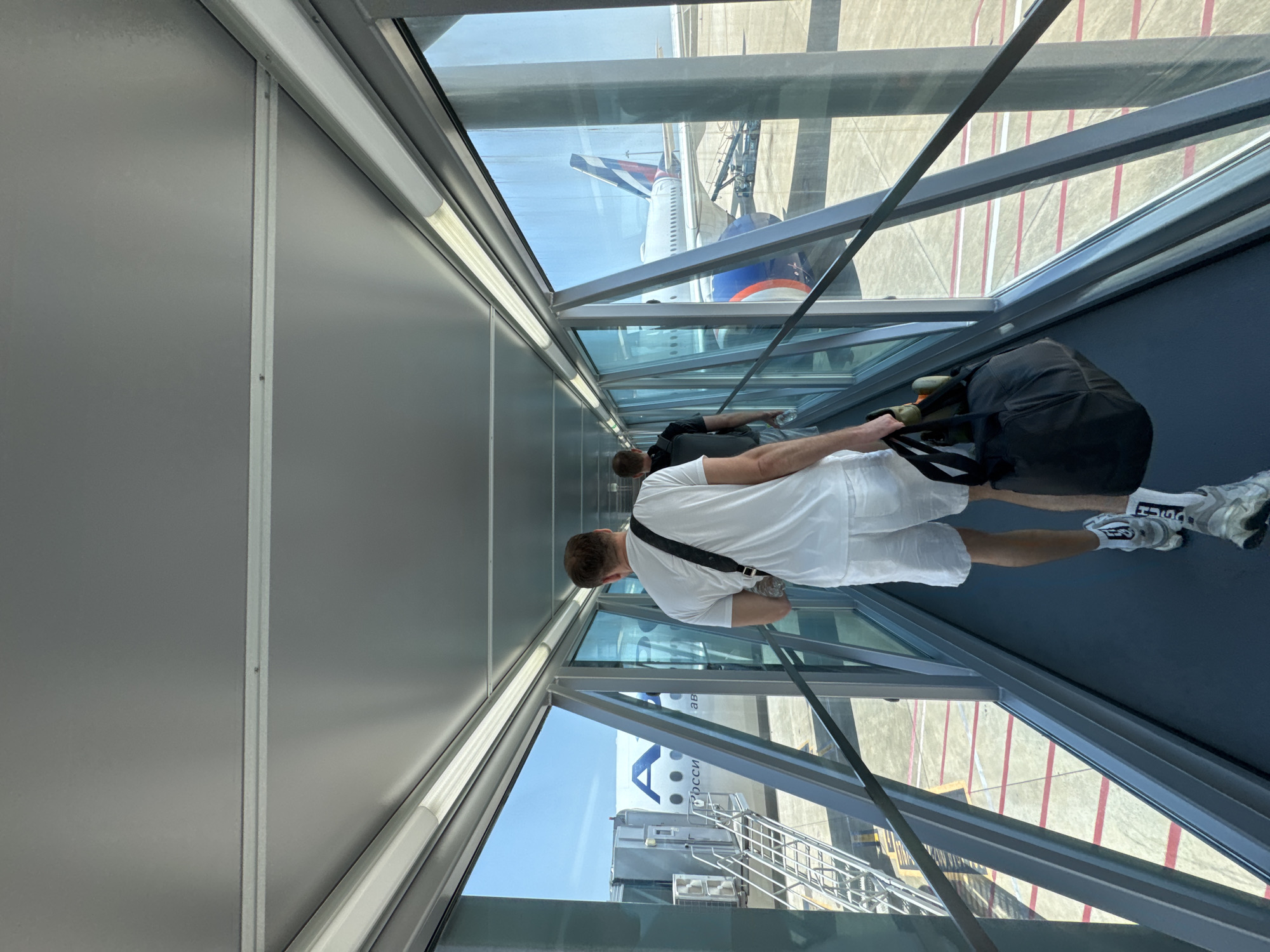 A person is walking through an enclosed airport walkway. Another person is visible ahead of them. The walkway has large windows on both sides, allowing a view of the outside. Through the windows, an airplane with a blue and red tail can be seen, as well as a boarding bridge connected to the plane. The walkway is made of metal and glass. The person walking is wearing white shorts, a white t-shirt, and white sneakers. They are carrying a black backpack.