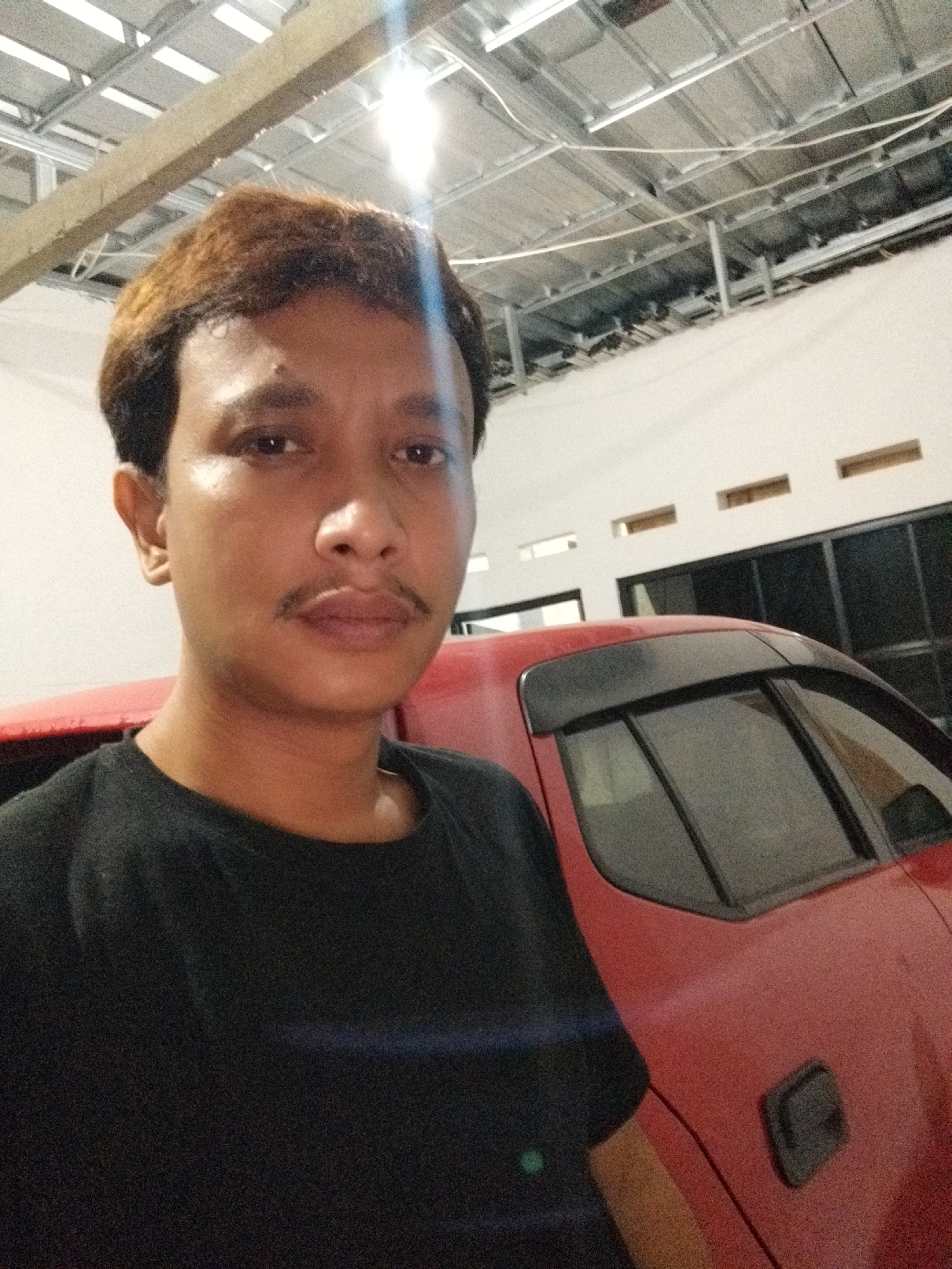 In Kaduhejo, Indonesia, a young man with brown hair and a mustache, wearing a black t-shirt, is taking a selfie in what appears to be a garage or workshop. Behind him, a red car is visible, with part of its side and rear window in frame. The background shows the unfinished ceiling of the structure, with exposed metal beams and some wiring. The lighting is dim, with a bright light source creating a glare on the ceiling and on the man's forehead. The overall impression is an informal, personal snapshot taken in an industrial or utilitarian space.