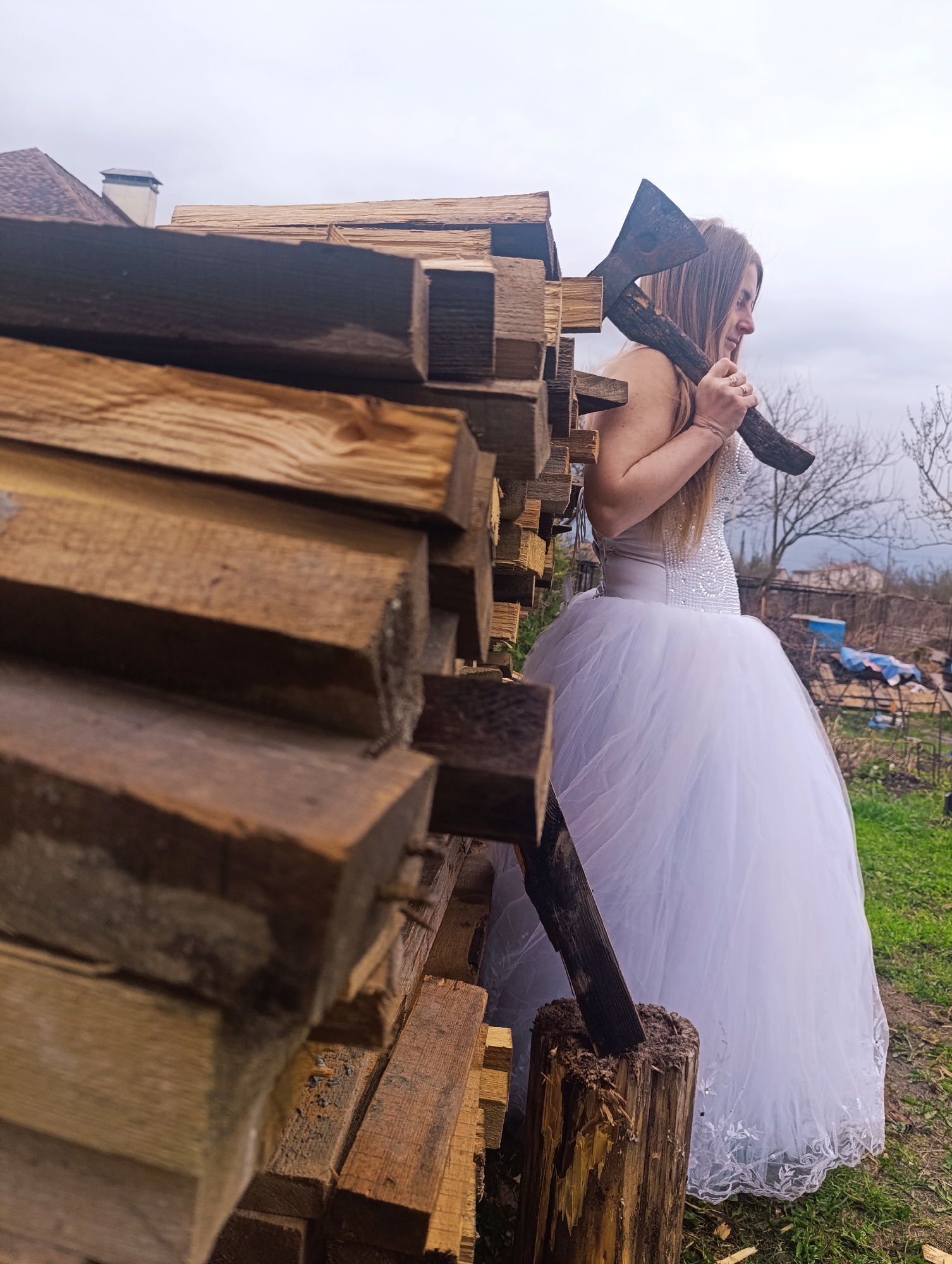 A woman in a white wedding dress stands in a rustic, outdoor setting, holding an axe over her shoulder. She is positioned near a stack of lumber and a tree stump, which has an axe in it. The environment appears to be a backyard or garden with grass, foliage, and a house in the background under a cloudy sky.