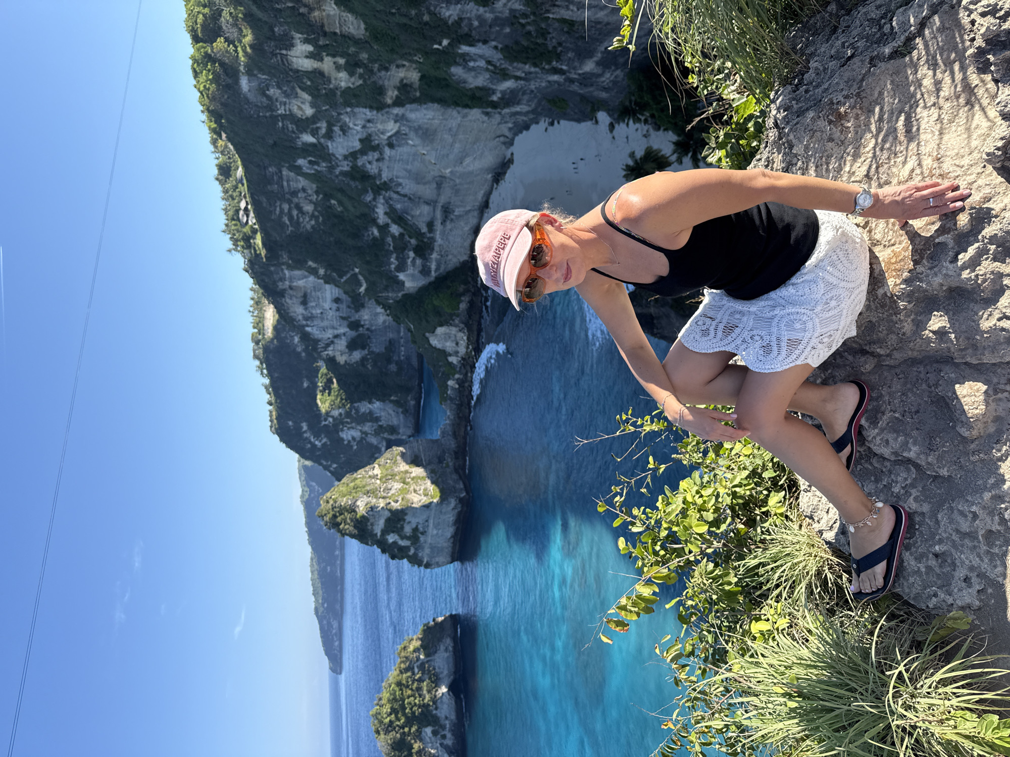 A woman wearing a black tank top, a white lace skirt, sunglasses, and a pink baseball cap sits on a rock on a cliff overlooking the ocean in Nusa Penida, Indonesia. The ocean is a vibrant turquoise, and dramatic cliffs with lush green vegetation rise from the water. A sandy beach is visible in the distance, nestled between the cliffs. The sky is clear and blue, with two faint lines that appear to be power lines or aircraft contrails. The woman is looking towards the camera, with her right arm resting on her knee. Her left arm is bent, with her hand resting on the rock beside her. She is wearing a watch on her left wrist and a ring on her left ring finger. Her feet are clad in dark flip-flops with red accents, and she has an anklet with small charms. The overall scene depicts a scenic and sunny day at a coastal location.