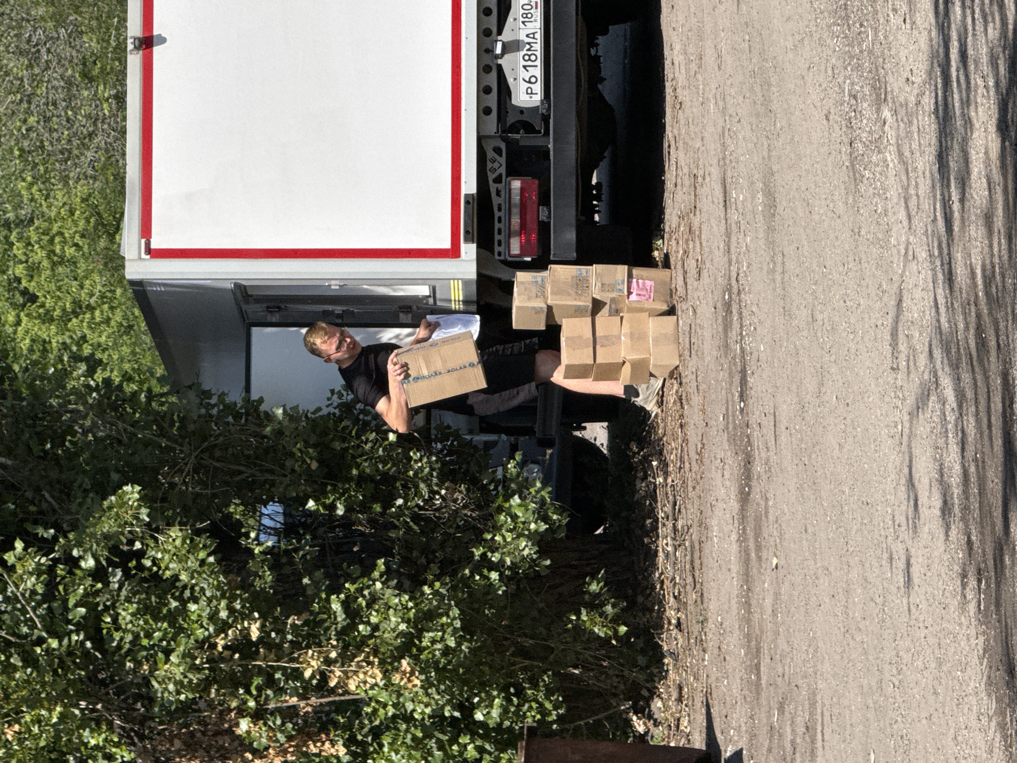 A young man with blond hair and glasses, wearing a black t-shirt and black shorts, is standing next to a white truck with a red trim. He is holding a cardboard box and appears to be in the process of unloading more boxes from the truck. Several cardboard boxes are stacked on the ground next to him. The truck has a license plate that reads "P618MA 180". The background shows green trees and foliage, and the ground is paved.