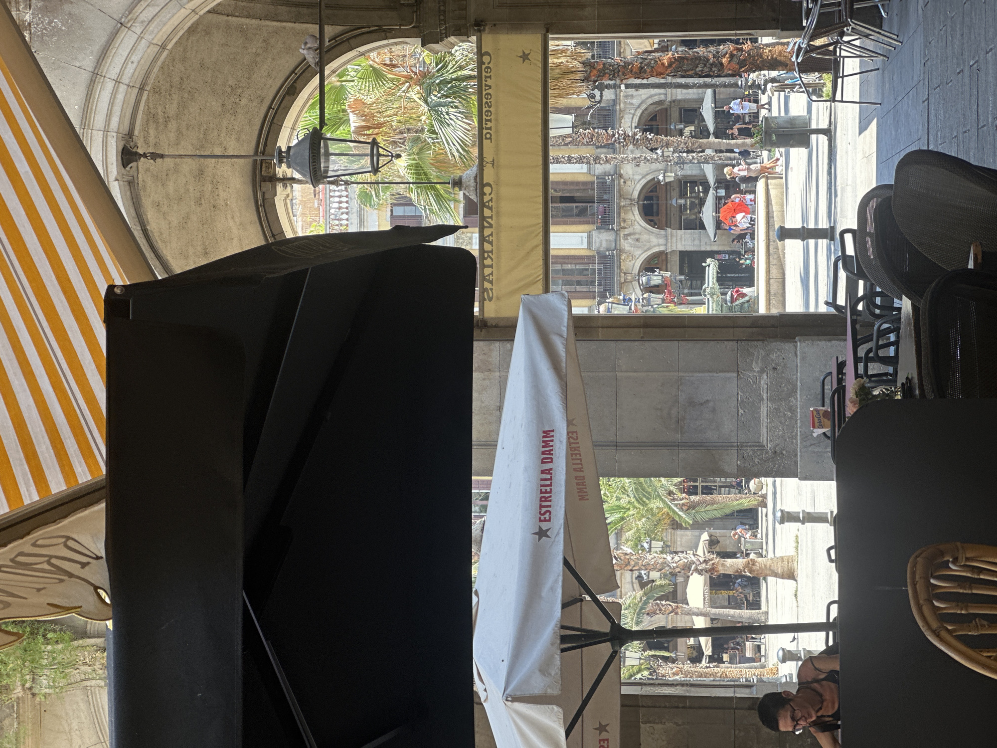 This image captures a street-level view of a covered outdoor seating area, likely a cafe or restaurant, in Barcelona, Spain. The foreground features a black awning and a white umbrella with the "ESTRELLA DAMM" logo. A woman with glasses is seated at a table in the lower left, looking down. Beyond the immediate seating area, an arched passageway opens to a sunlit public square. Palm trees are visible throughout the square, along with more covered seating areas and people walking. In the background, the facade of a building with arched windows and balconies is present. The overall atmosphere suggests a warm, sunny day.