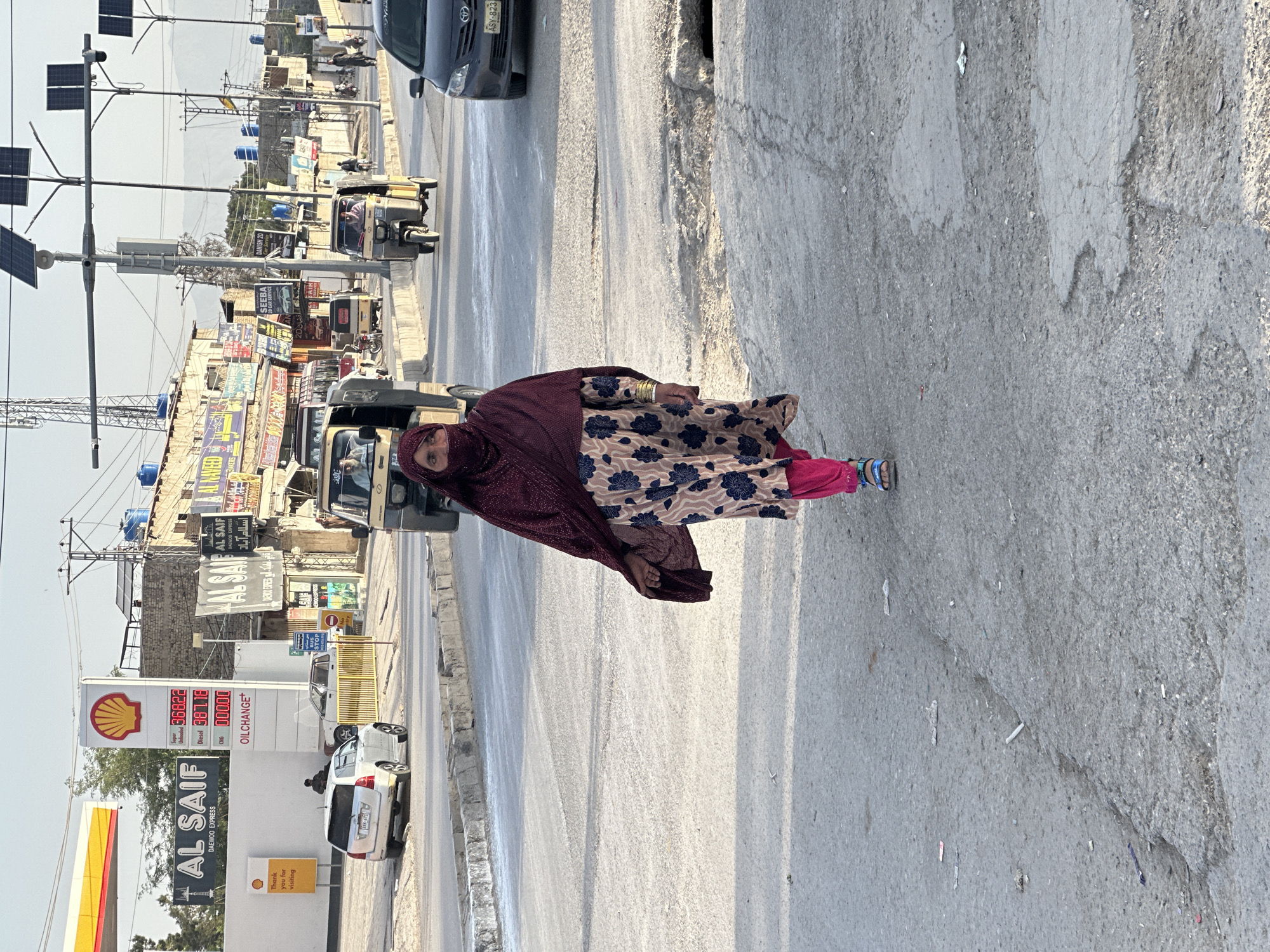 This image, captured in Quetta City Tehsil, Pakistan, presents a busy street scene under bright daylight. The main subject is a woman standing on the right side of the cracked asphalt road, facing slightly towards the camera. She is dressed in a maroon head covering, a long dress with a floral pattern (blue flowers on a light background), pink trousers, and bangles on her right wrist, suggesting traditional attire.
The road is active with various vehicles. A dark-colored car with the license plate "ASY-823" is seen driving away in the mid-ground. Several auto-rickshaws, a common sight in the region, are also visible, one with a passenger. Along the left side of the road, buildings house multiple shops and businesses, adorned with numerous signs, many written in Urdu.
A prominent Shell petrol station, identified as "AL SAIF DAEWOO EXPRESS," is visible on the left. Its signage displays current fuel prices: Super Unleaded at 368.22, Diesel at 387.18, and CNG at 00.00 (likely unavailable or not priced), along with "OILCHANGE+" and a "Thank you for visiting" message. Utility poles equipped with solar panels line the street, carrying a network of electrical wires. Yellow roadside barriers and blue water tanks on building rooftops are also notable. In the hazy distance, mountains or hills are faintly visible, contributing to the geographic context. The clear sky indicates pleasant weather, and the overall scene depicts a typical, bustling urban commercial area.