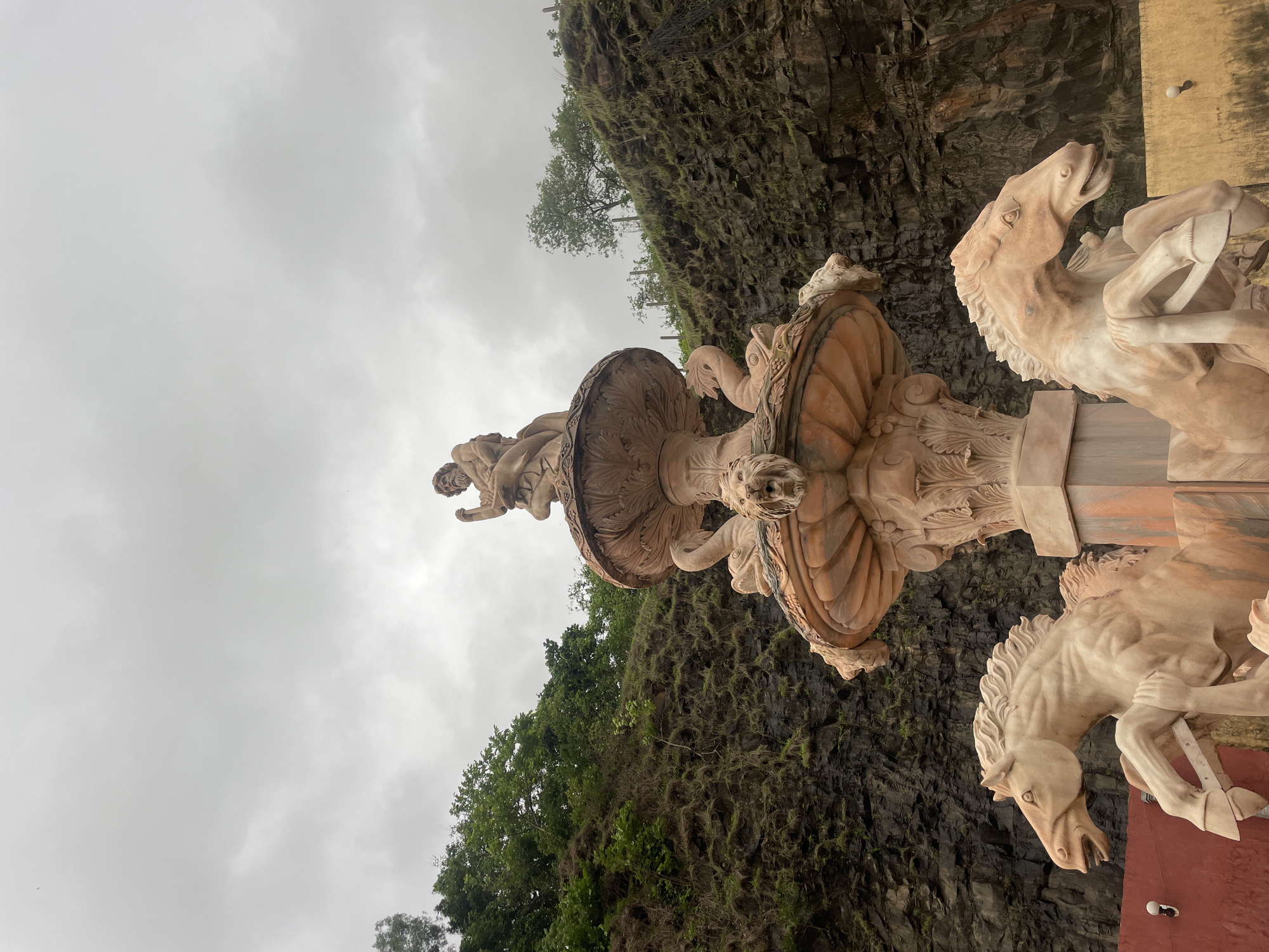 The image shows a stone fountain sculpture of horses and a statue of a person, set against a backdrop of a rocky cliff face with vegetation and trees. The sculpture appears to be made of light-colored stone, possibly marble or sandstone, and features intricate detailing. Three horses are prominently depicted, with one in the foreground appearing to be rearing up. A human figure, possibly a god or mythological character, is visible on top of the fountain structure.
The scene is outdoors, with a cloudy sky indicating overcast weather. The rocky cliff is dark and textured, with some greenery clinging to its surface, suggesting a natural or semi-natural environment. The presence of trees further contributes to the natural setting. The composition is dynamic, with the figures and the cliff face creating a sense of grandeur.
The time of day appears to be daytime, given the ambient light, but the cloudy conditions diffuse the sunlight. There are no visible signs of text in the image. The overall mood is one of artistic grandeur and natural beauty, with the sculpture being a focal point against the rugged backdrop. The location context provided is Mumbai, India, which might suggest the sculpture is part of a park, garden, or historical site within the city.