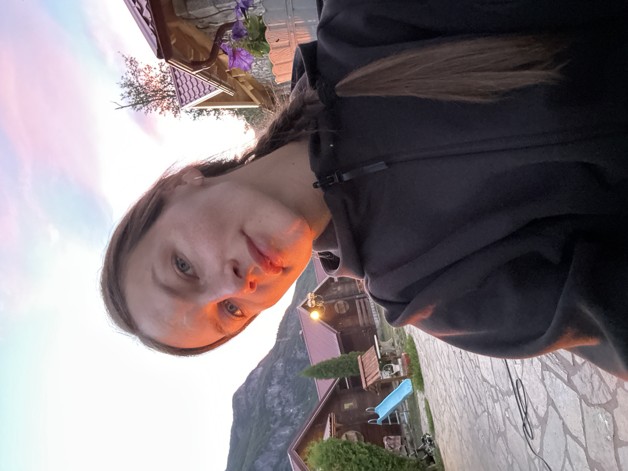 The image shows a woman taking a selfie outdoors in the city of Gusinje, Montenegro. The woman is wearing a dark hooded jacket and has her hair in a braid. Her face is partially illuminated by the setting sun, creating a warm glow. In the background, there are several wooden houses with red roofs, a blue slide, and a mountainous landscape. The sky is a mix of blue, pink, and orange hues, indicating that it is either sunrise or sunset. The ground is paved with stone tiles, and a black cable is visible on the ground. The overall scene suggests a peaceful, rural setting during golden hour.