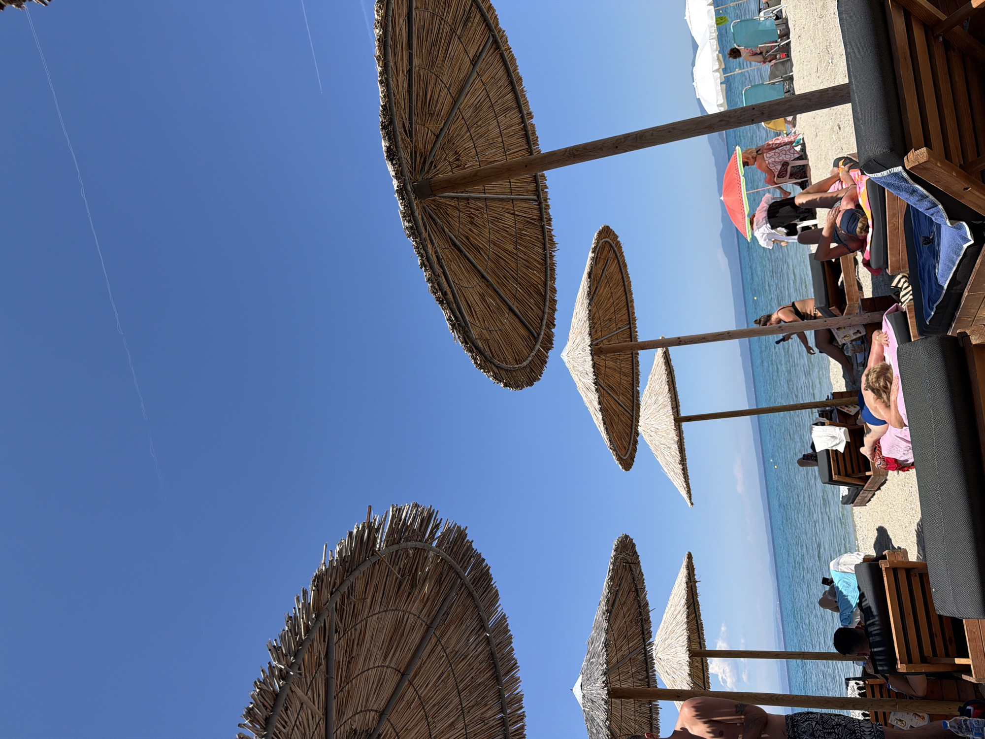 The image captures a sunny day at a beach in the Pallini Municipal Unit, Greece. Several straw umbrellas provide shade over lounge chairs occupied by people. The blue Ionian Sea stretches to the horizon. Two contrails streak across the clear blue sky.