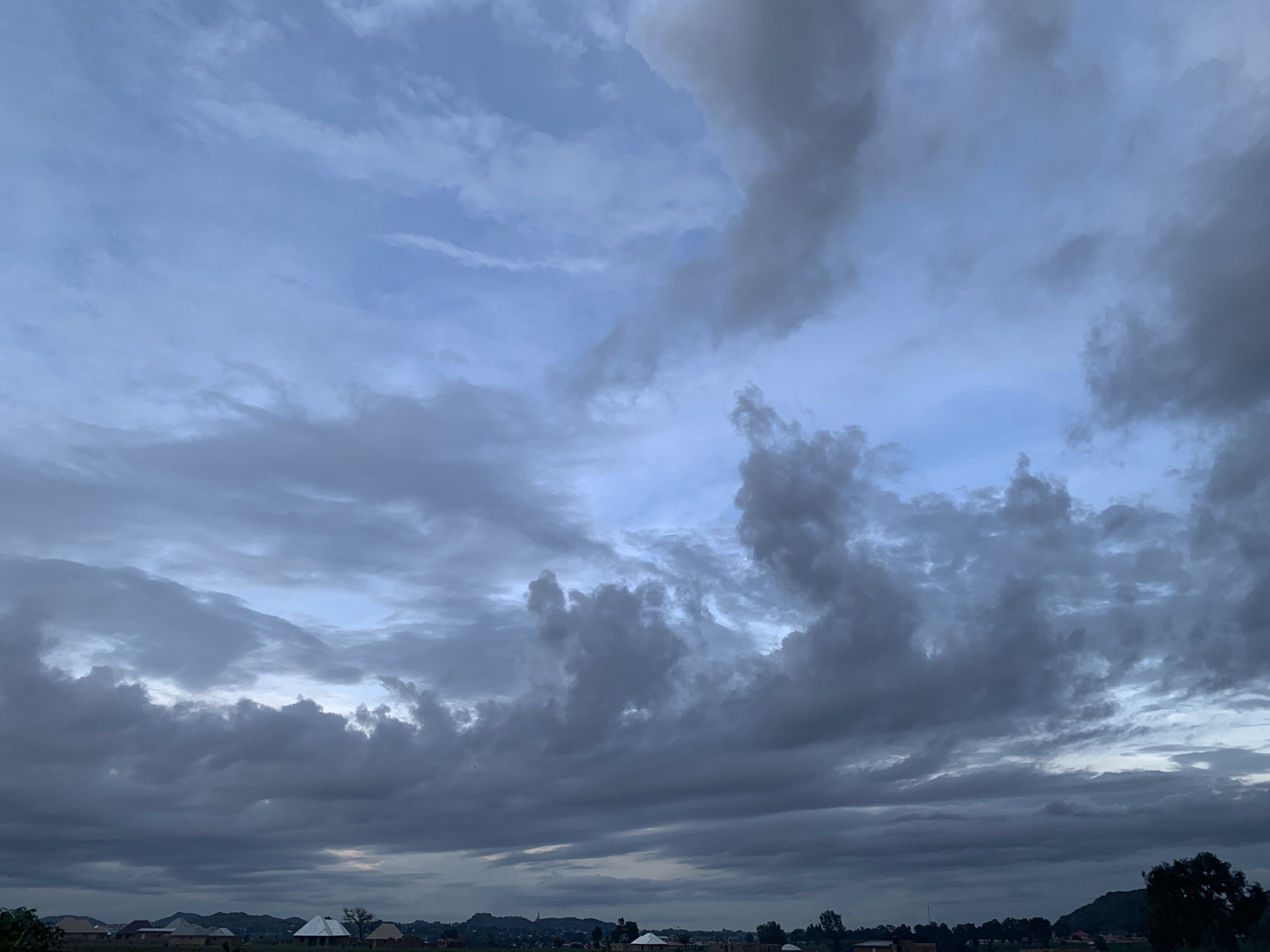 The image captures a dramatic sky filled with a mix of light and dark, textured clouds, suggesting an overcast or transitioning weather condition. The sky dominates the frame, with lighter blue hues visible in patches, indicating breaks in the cloud cover or perhaps the approaching dawn or dusk. The clouds themselves are varied in form and density, with some appearing wispy and others thick and heavy, casting a somber or brooding mood.
Below the expansive sky, a landscape unfolds with hints of human settlement. A line of buildings, characterized by simple roofs, are visible along the horizon, suggesting a rural or semi-urban setting. Trees and vegetation are interspersed, and rolling hills or mountains form a backdrop in the distance. The overall impression is one of a quiet, possibly developing area.
The time of day is suggested by the quality of light and the cloud formations; it appears to be either early morning or late afternoon, a period often associated with diffused light and dramatic skies. The weather is clearly a significant element, with the clouds indicating potential for rain or having recently experienced it. The location in Kuru Karama, Nigeria, provides a geographical context for the vegetation and architectural styles that might be present. No text is visible in the image.