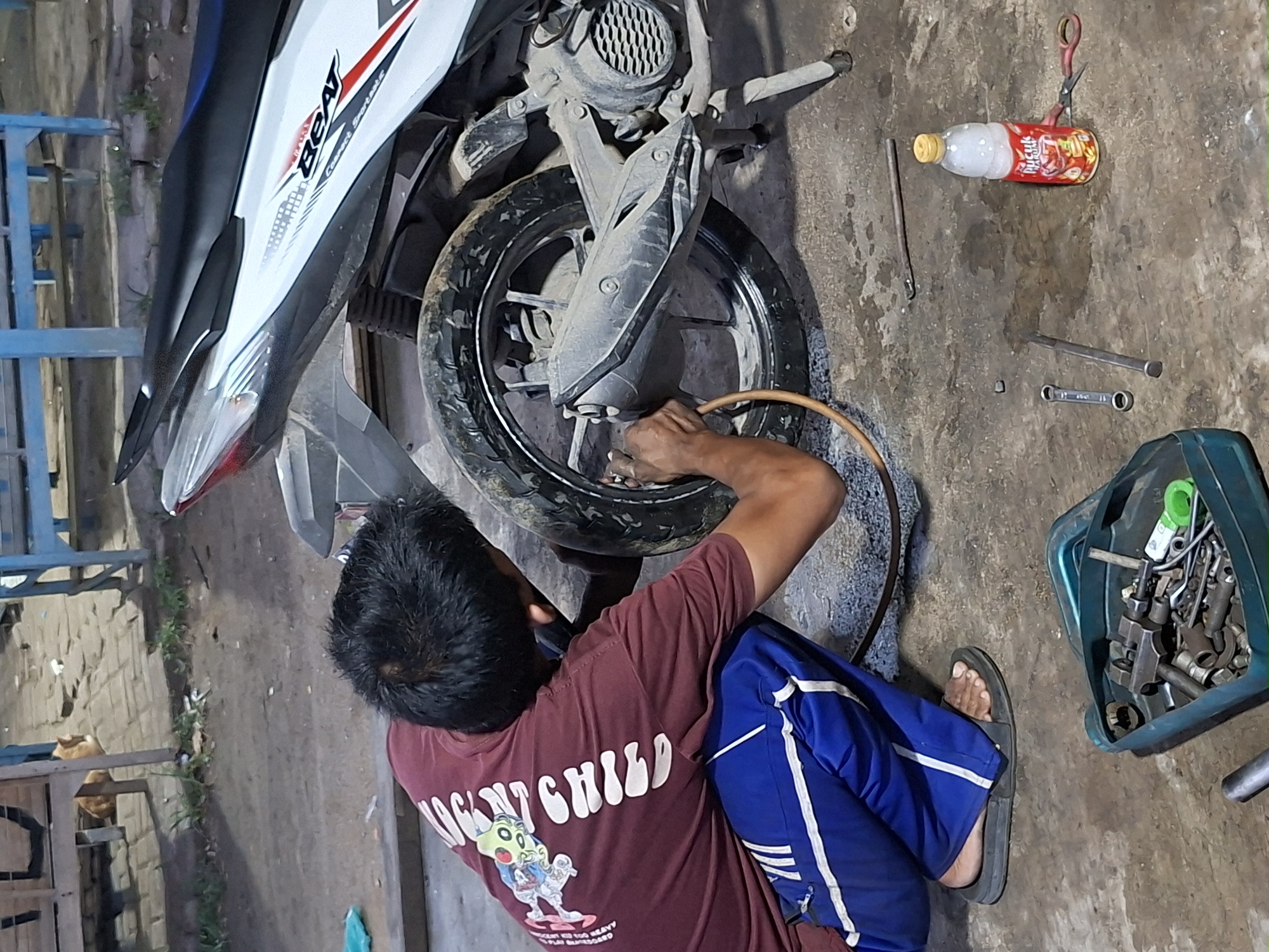 A person is crouched down, inflating the rear tire of a white and black Honda Beat motorcycle. The person is wearing a maroon t-shirt with a graphic and text on the back, and blue athletic pants with white stripes. They are using an air hose connected to a portable compressor. In the foreground, a teal toolbox is filled with various wrenches and tools. A bottle of soda and a pair of scissors are also on the ground nearby. The scene appears to be an outdoor workshop or repair area, possibly in Lhokseumawe, Indonesia, given the context provided. The ground is concrete or dirt, and there are glimpses of wooden structures and blue metal framing in the background, suggestive of a street-side establishment.