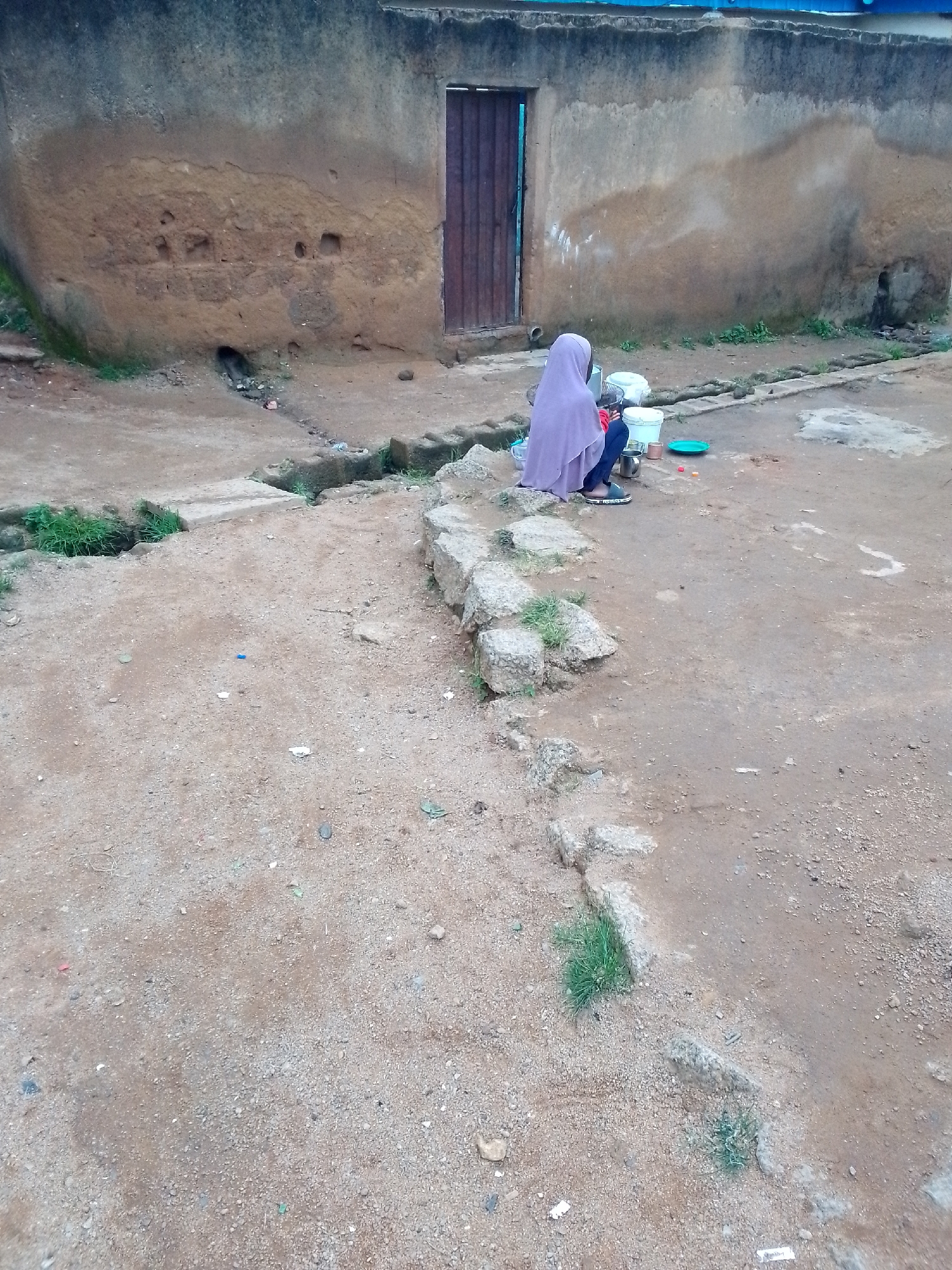 A medium shot captures a person sitting on a low stone wall in front of a building with a rough, textured wall. The person, wearing a purple hijab and blue pants, is facing away from the camera, looking down at items on the ground. To the right of the person are two white buckets, a green plate, and a red object, suggesting they are engaged in an activity such as washing or preparing something. The ground is dirt and sand, with scattered small stones and patches of grass. A narrow, shallow trench runs horizontally across the middle ground of the image, bordered by more stones and vegetation. The building behind has a dark wooden door and appears to be made of adobe or similar material. The overall scene depicts an outdoor, possibly rural or less developed, urban setting.