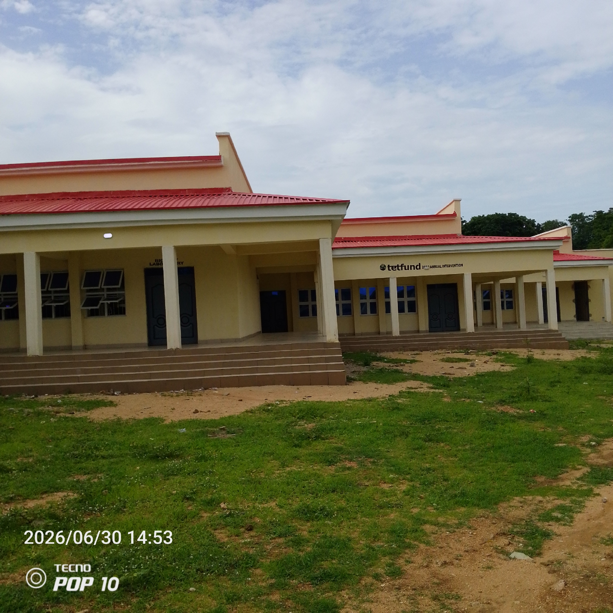 This is a full, eye-level, outdoor shot of a light yellow, single-story building complex with red roofs, located in Jalingo, Nigeria. The building appears to be a public institution, possibly an educational or administrative facility. A sign on the right side of the building reads "tetfund 2022 ANNUAL INTERVENTION". The building features several entrances with dark blue doors and windows with blue panes. White columns support the roof over a walkway. The building is surrounded by green grass and dirt patches. In the foreground, on the grass, is a timestamp indicating "2026/06/30 14:53" and a Tecno POP 10 logo. The sky is cloudy. There are no people visible in the image.