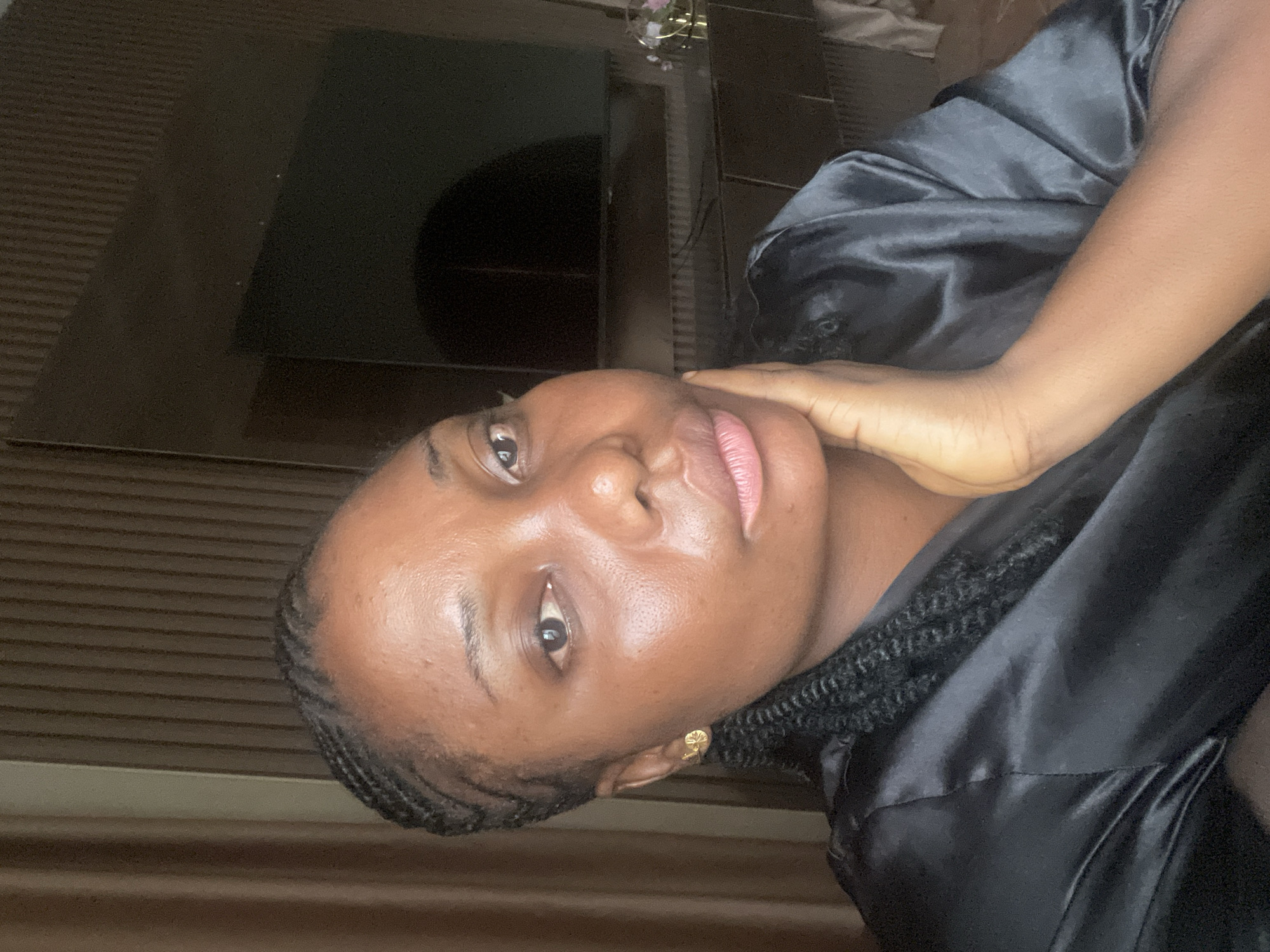 The image is a selfie-style portrait of a woman with dark skin, wearing a black satin robe. Her hair is styled in braids. Her left hand rests on her neck. An earring is visible in her right ear. The background consists of a dark surface, possibly a television screen, and horizontal wooden panels. It is not possible to confirm the location as Jos, Nigeria from the image.