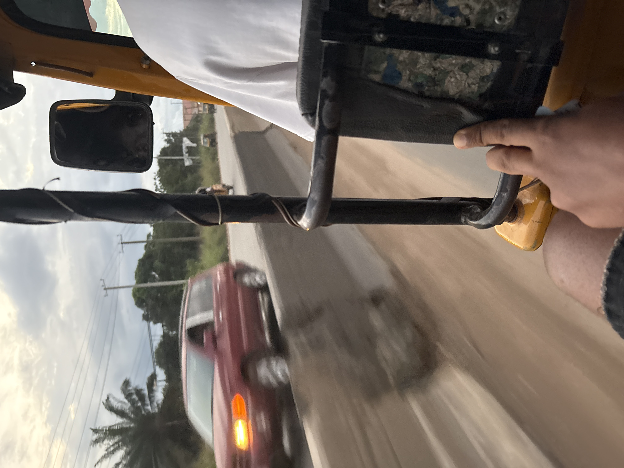 The image captures a street scene in Onitsha, Nigeria, from the perspective of someone inside a motorized tricycle, commonly known as an "Okada" or "Keke Napep." The foreground shows a hand resting on the edge of the tricycle's seat, with a person's leg visible below. The tricycle itself is yellow, and parts of its frame and a side mirror are visible.
In the mid-ground, a reddish-brown SUV is driving on the road, its headlights on, indicating it might be dusk or a shaded area. The vehicle appears to be in motion, as are other vehicles in the distance. The road is lined with a concrete barrier, suggesting it might be a highway or a major road. Trees and utility poles with overhead wires are visible along the side of the road, typical of an urban or semi-urban environment in Nigeria. The sky is cloudy, with shades of grey and white, hinting at possible impending rain or the end of the day. The overall impression is a snapshot of everyday life and movement in Onitsha.