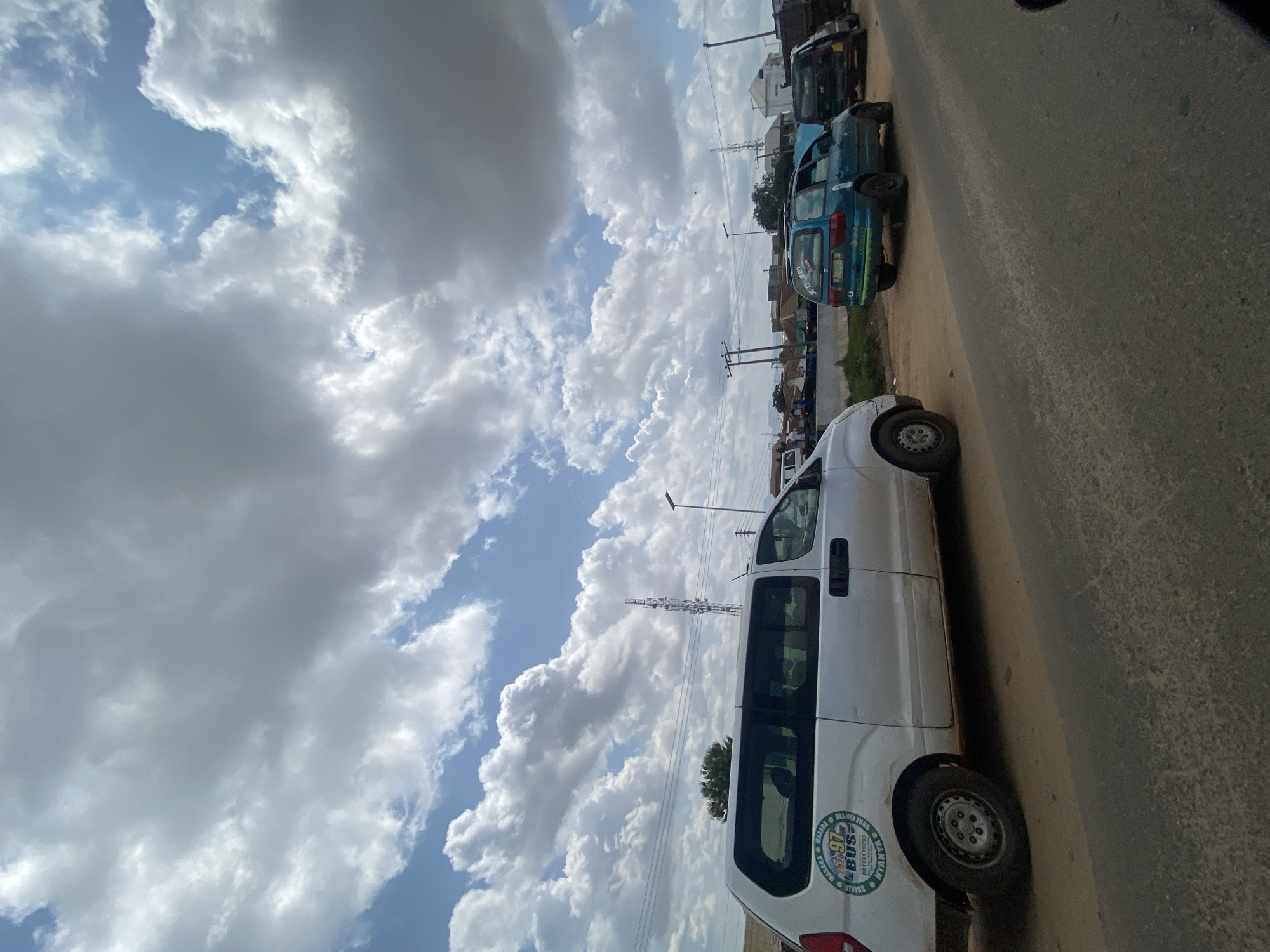 This is a medium-angle, eye-level shot of a street scene in Ado, Nigeria. In the foreground, a white van is parked on a dirt shoulder next to a paved road. The van has a circular decal on its side featuring the number "97 BUS" and text that reads "DUTE, BANANA, MAJARA, NYANYA." Further down the road, a teal-colored car with its hood up is stopped. Behind the teal car, a black SUV is also stopped, and its passenger door is open. In the background, there are several buildings, utility poles, and a cell tower. The sky is filled with large, dramatic clouds. The overall scene suggests a busy roadside environment, possibly near a transport hub or market, with vehicles stopped for various reasons.