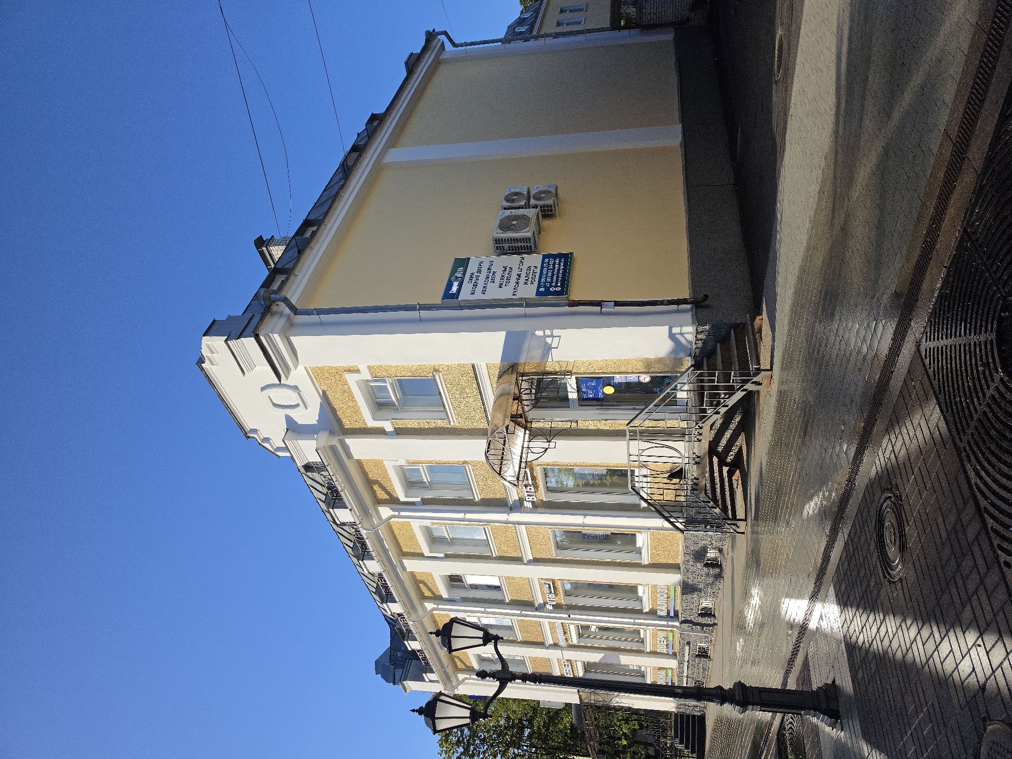 This is a full, low-angle, outdoor, eye-level, medium shot of a two-story building in Gatchina, Russia. The building is light yellow with white trim and a textured facade. It has a gray, sloped roof with dormer windows. The ground floor appears to be commercial, with several windows displaying signage. A black lamppost with two lights stands to the left of the building. The sidewalk in front of the building is paved with dark gray bricks and is wet, reflecting the sunlight. There is a set of metal stairs leading up to an entrance. On the side of the building, there are multiple air conditioning units installed. A blue and white sign on the building lists services such as "windows, exterior doors, interior doors, stretch ceilings, roller shutters, blinds." The sky is a clear, bright blue. The scene is set in an urban environment, with shadows cast by the building and lamppost across the wet sidewalk.