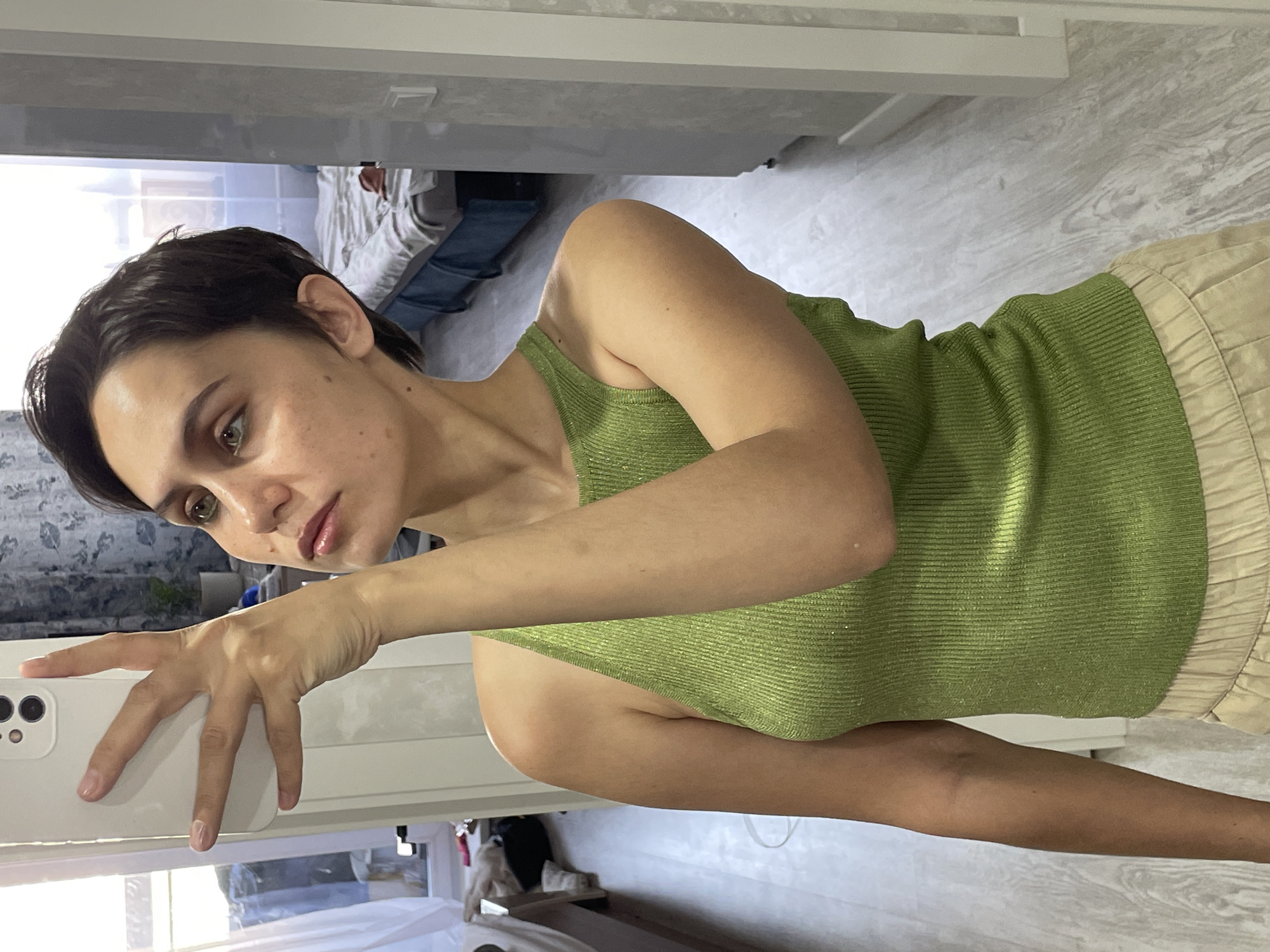A woman is posing for a selfie, holding a white smartphone with a triple-lens camera in her left hand. She is wearing a green tank top and beige bottoms. The background shows an interior scene with a bed visible through a doorway and a window with curtains. The floor is light-colored. The location is likely Izhevsk, Russia, based on contextual knowledge.