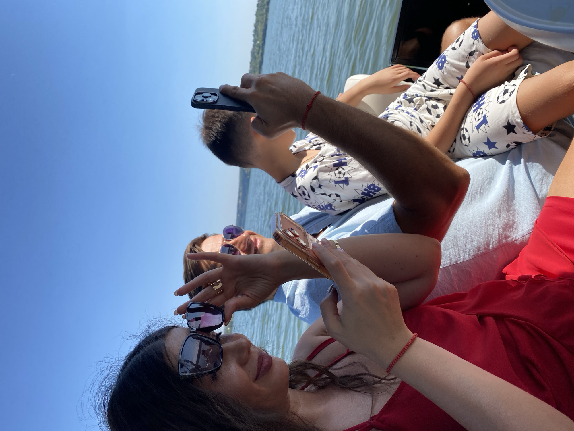 In Molovata, Moldova, a group of three individuals, two adults and one child, are enjoying a sunny day on a boat. The woman in the foreground, wearing a red dress and sunglasses, is looking down at her phone. Next to her, a man in a blue shirt also appears to be using his phone. In the background, a child in a white t-shirt with a soccer ball print is seated. The boat is on a body of water, with the lush green banks of the river visible in the distance under a clear blue sky.