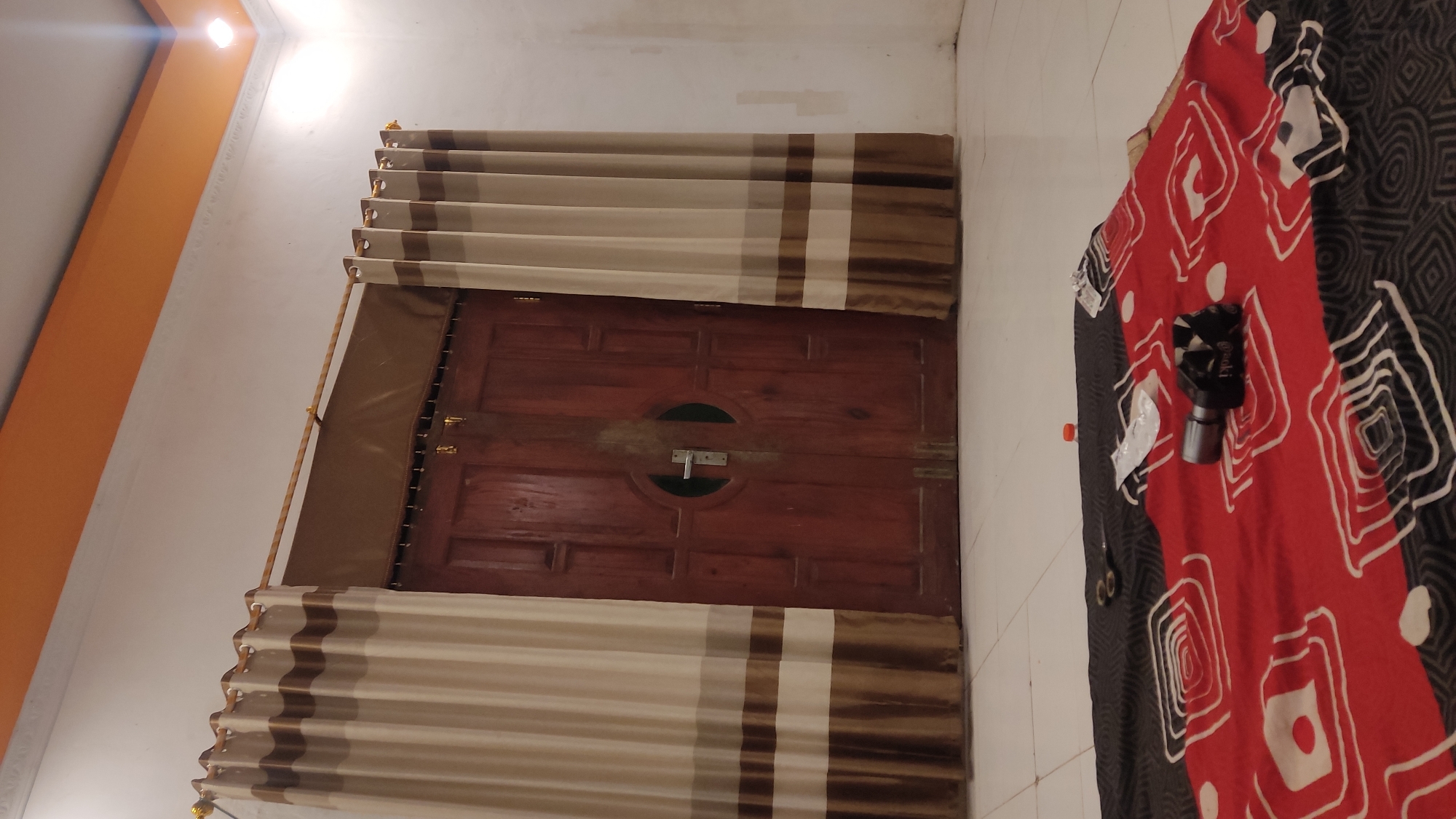 The image depicts a room in Ngadiharjo, Indonesia, featuring a pair of wooden doors with a circular glass inlay and a silver handle. The doors are flanked by beige and brown striped curtains. In the foreground, a patterned blanket with red, black, and white designs is spread out on a tiled floor. On the blanket, there are several objects including a pair of scissors, a small orange bottle cap, and what appears to be a small camera or electronic device. The room has white walls and an orange ceiling trim. A light fixture is visible in the upper right corner.