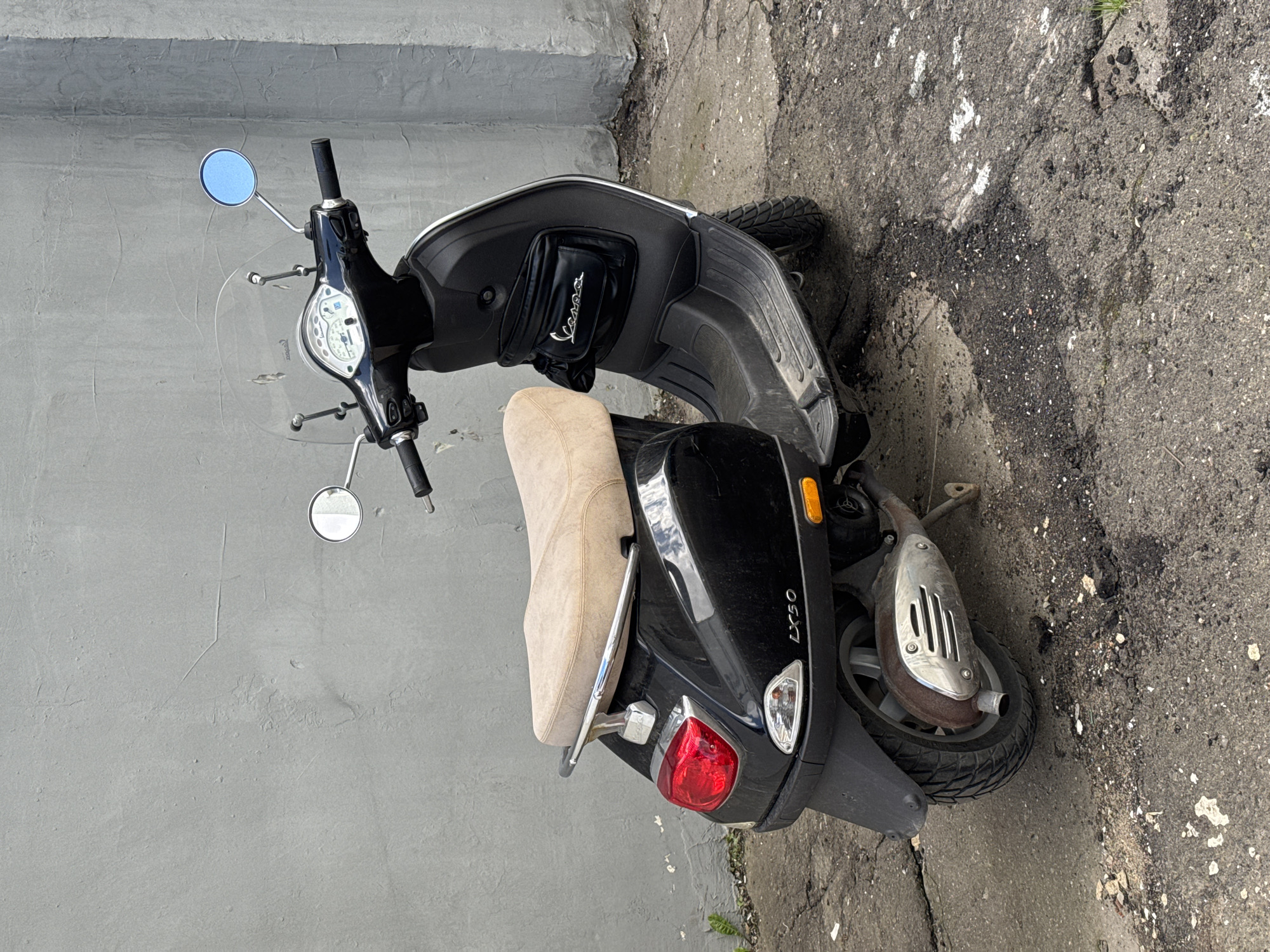 A black Vespa LX50 scooter is parked on a cracked asphalt surface next to a grey concrete wall. The scooter has a beige seat, a clear windshield, and a black bag with the "Vespa" logo attached to the front. The scooter's rear light is red, and there is an orange turn signal visible. The ground is uneven and appears to be in disrepair, with patches of exposed earth and debris. There are no people in the image. The scene is likely in an urban or industrial area, given the concrete wall and the state of the pavement.