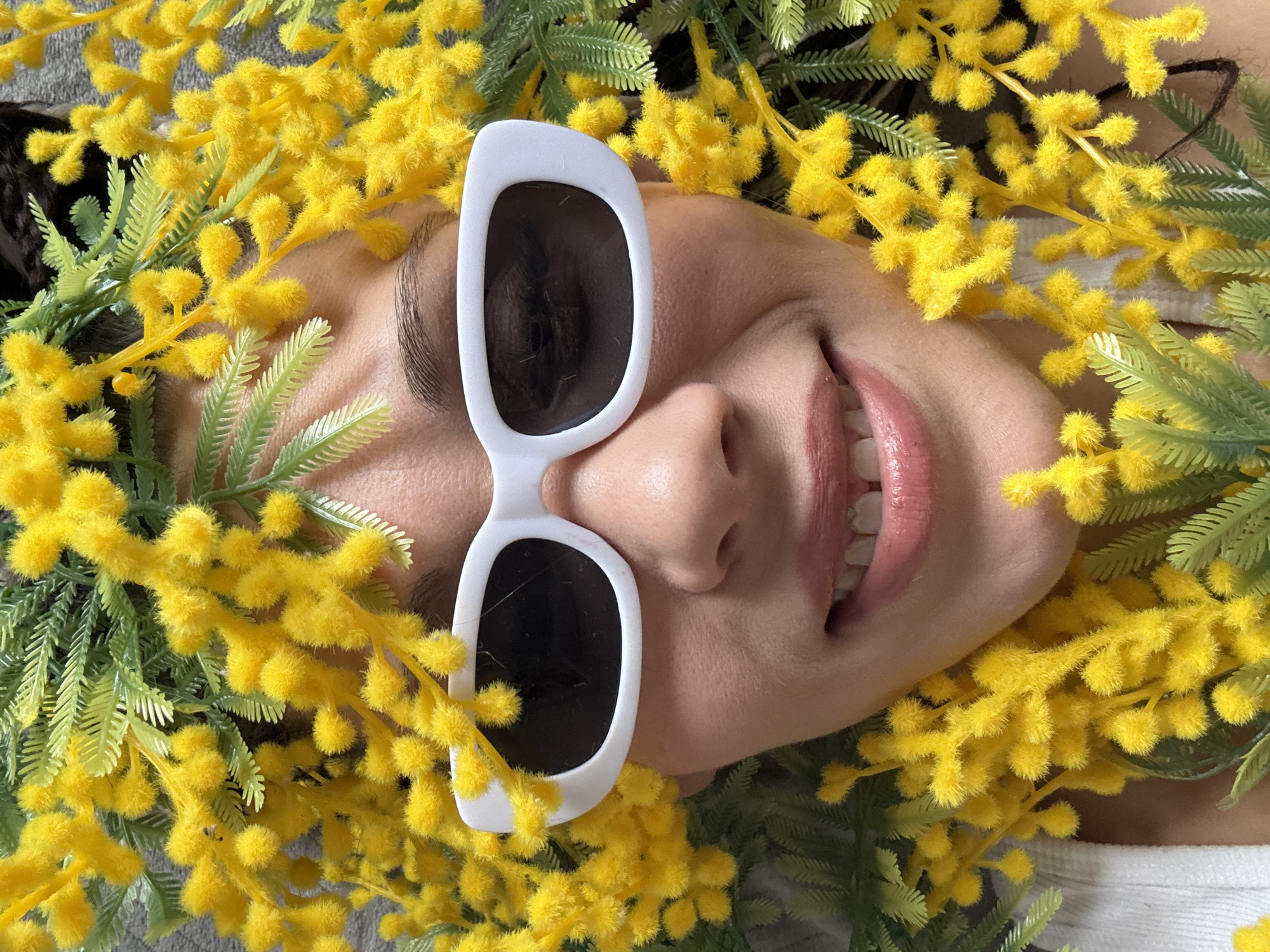 A woman lies amidst sprigs of yellow mimosa flowers and green foliage, wearing white-framed sunglasses with dark lenses. Her face is oriented at an angle, with her nose and mouth visible. A portion of her hair is visible in the upper-left corner. The background is blurred. There is no information to suggest the location in the city of Bor, Russia.