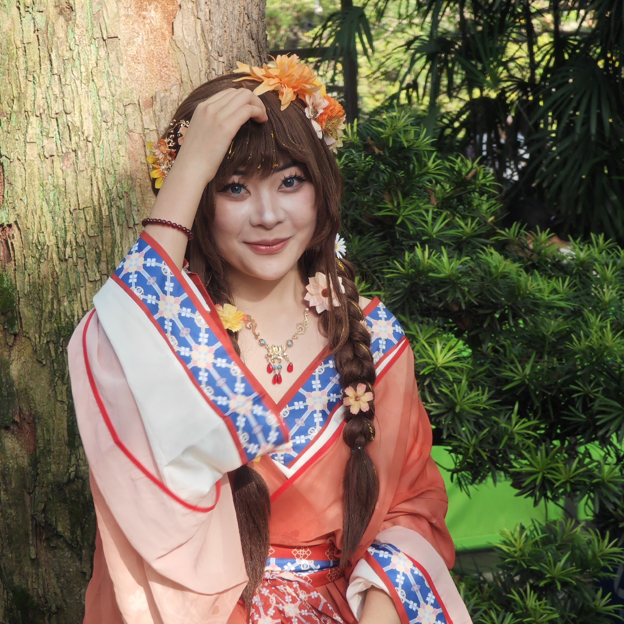 A woman in traditional Hanfu clothing stands next to a tree in an outdoor setting. She wears a floral crown, has braided hair adorned with flowers, and is holding her hair with her right hand. She is also wearing a necklace. The environment includes greenery like bushes and possibly palm trees, suggesting a garden or park. The Yuzhong District, China, is not discernible from this image alone.