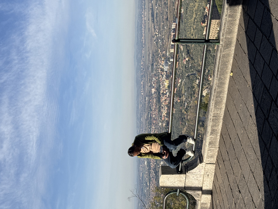 The scene depicts an individual seated on a metal railing at a panoramic overlook in Tivoli, Italy, under a clear blue sky with sparse, wispy clouds. The foreground consists of a dark grey stone-paved terrace leading to a light-colored concrete balustrade. A dark grey metal railing, comprising two horizontal bars, is affixed to the balustrade.
A single male subject, with dark hair, is positioned centrally, sitting on the top horizontal bar of the railing, facing generally towards the right (east or southeast). He wears a dark olive-green hooded jacket over a light tan t-shirt, dark blue jeans, and white athletic shoes with dark soles and a visible dark logo (appears to be a Nike swoosh). His head is tilted downwards, focused on a dark-colored smartphone held in both hands, suggesting engagement with the device.
Beyond the railing, a vast, hazy panoramic view unfolds. This includes a dense urban area with numerous buildings of varying sizes and roof colors (predominantly red and orange) interspersed with green areas, likely cultivated land such as olive groves. Distant hills or mountains are visible on the horizon, partially obscured by atmospheric haze. A large, multi-story building with a light brown facade is prominent in the lower right section of the panoramic view. The lighting suggests bright sunlight, casting distinct shadows from the railing and the individual onto the paved terrace. No other people are visible.