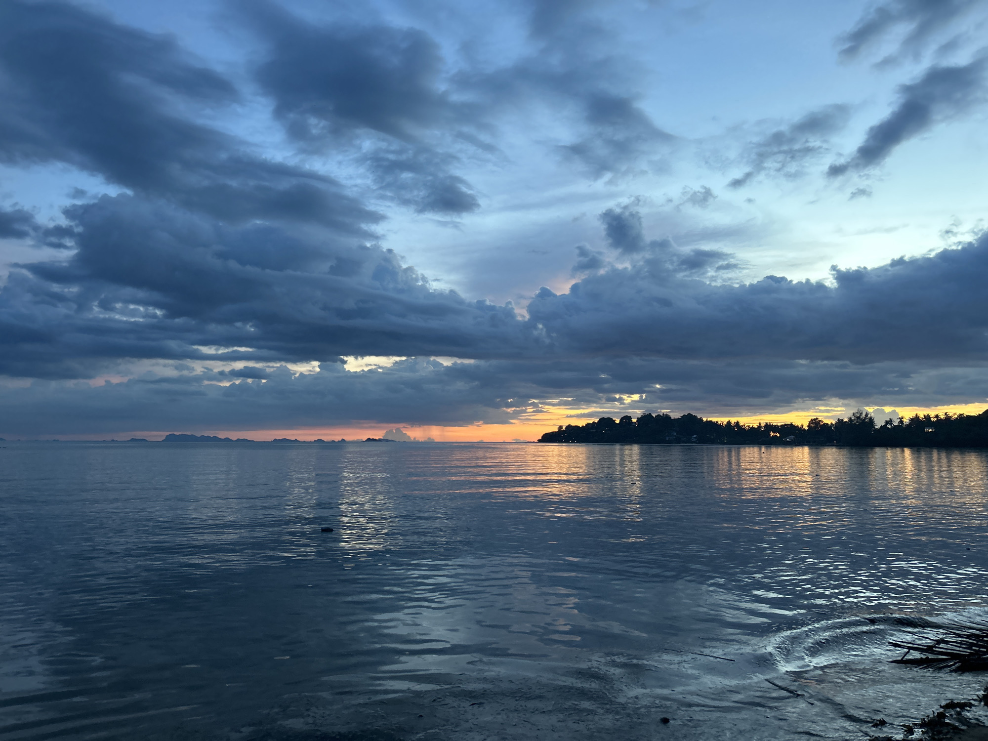 The image depicts a serene seascape at dusk, with a body of water, likely the Gulf of Thailand, in the foreground and middle ground. The water is calm, with gentle ripples reflecting the dimming light. In the distance, a silhouetted tree line marks the shore of what appears to be an island, possibly Koh Phangan itself, with a few scattered lights suggesting a settlement. The sky is a dramatic canvas of dark, moody clouds interspersed with patches of soft blue and hints of orange and yellow from the setting sun. The overall impression is one of tranquility and the impending end of a day in a coastal region of Thailand.