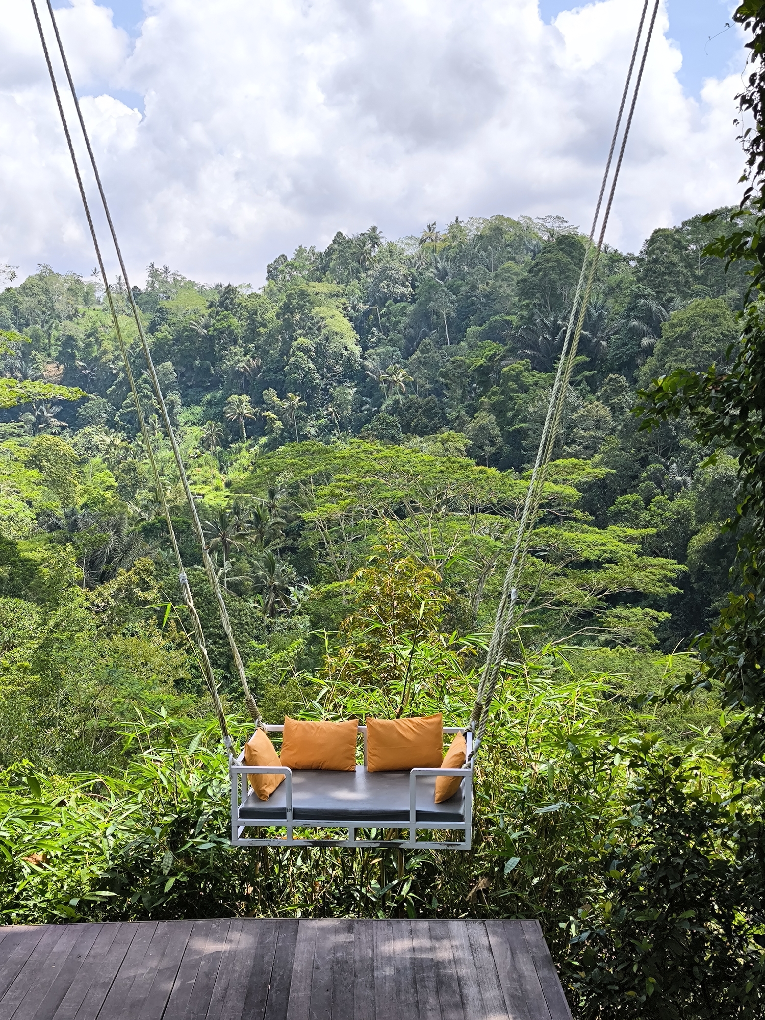 The image depicts a swing suspended over a lush, green valley. The swing has a gray platform with three orange pillows. The platform is supported by two sets of ropes that angle upwards. Below the swing is a wooden deck. The background features a dense forest with trees of varying heights and shades of green. The sky above is cloudy with patches of blue. This scene is located in Mekarsari, Indonesia.