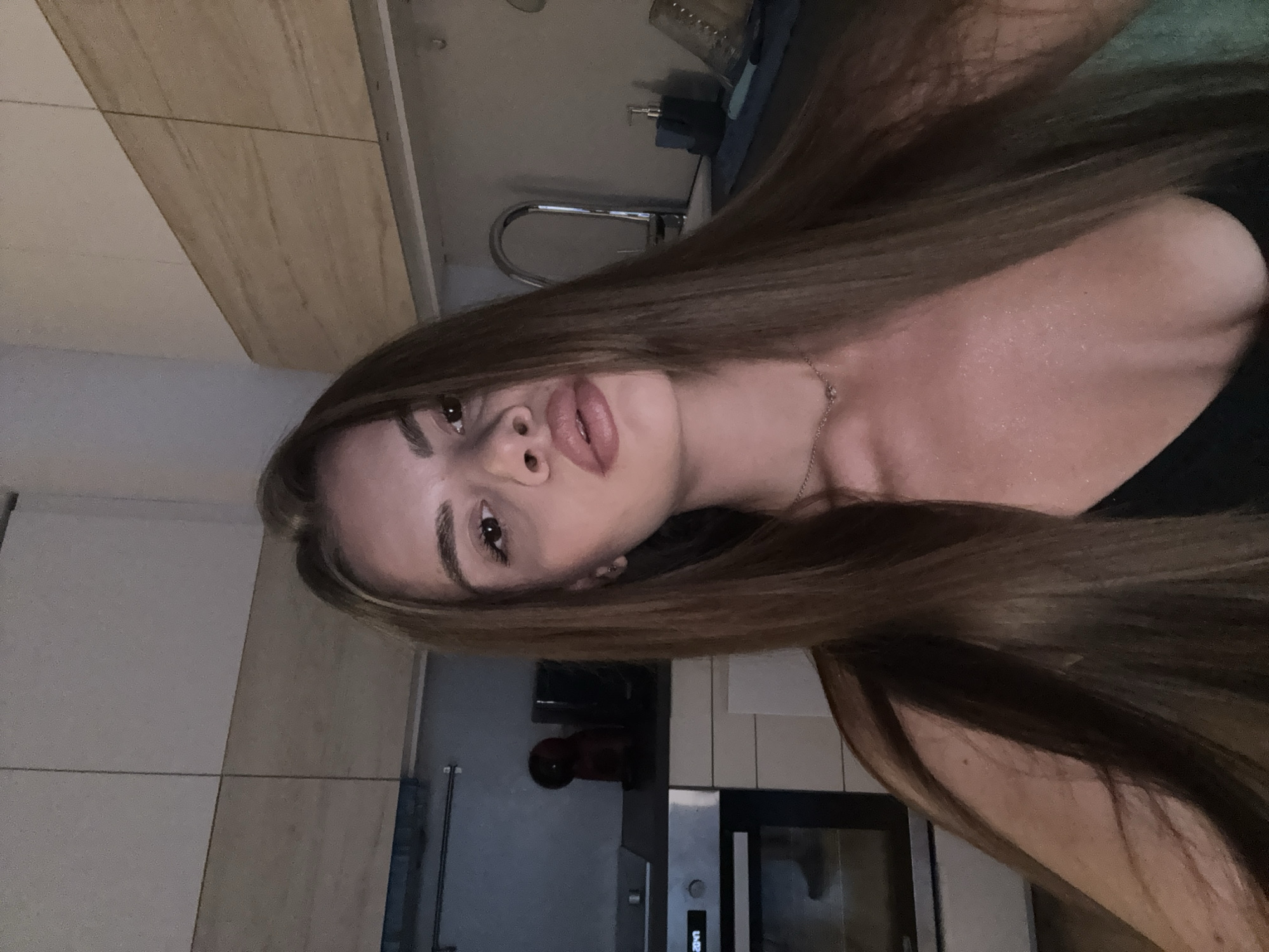 An indoor portrait-style selfie captures a young woman with long, straight, medium-brown hair. She is looking directly at the camera with a posed expression, her full lips slightly parted and her eyebrows defined. A delicate silver necklace is visible around her neck, and she wears a dark top. The lighting is dim, suggesting an evening setting.
The background elements indicate a kitchen environment. To the left of her head, light-colored walls or cabinetry and wooden paneling are visible. Behind her, a chrome kitchen faucet and sink are discernible, along with a soap dispenser and a blue item, possibly a sponge or cloth. Further to her right, a modern oven or microwave unit is integrated into the cabinetry, displaying "20:42" on its digital clock, confirming the time as 8:42 PM. White rectangular tiles form a backsplash behind the appliance. The setting appears to be a contemporary home interior, located in Plovdiv, Bulgaria.