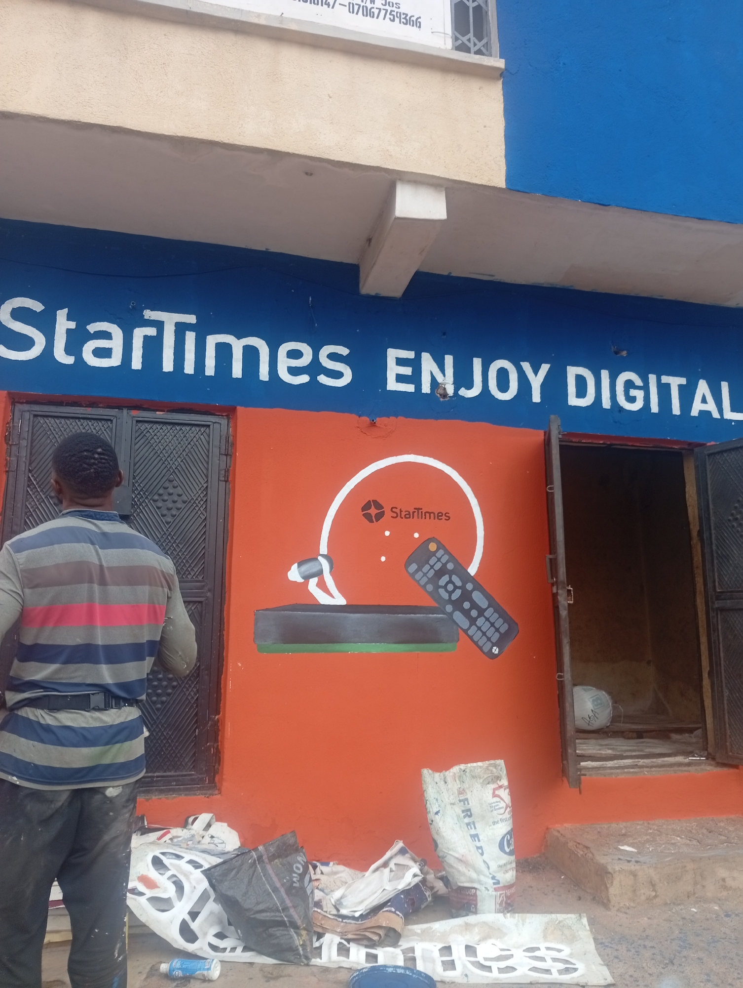 In Jos, Nigeria, a man is seen from behind wearing a horizontally striped shirt and dark pants, standing in front of a building. The building's facade is painted a vibrant orange, with a blue band across the top featuring white lettering that reads "StarTimes ENJOY DIGITAL". A mural on the orange wall depicts the StarTimes logo, a satellite dish, and a remote control, suggesting a business related to satellite television services. The man appears to be a painter, as indicated by the paint splatters on his pants and the surrounding area. In front of the building, on the ground, are various discarded materials, including plastic bags, sacks, and what looks like a partially painted sign. A doorway to the right is partially open, revealing the interior of the establishment.