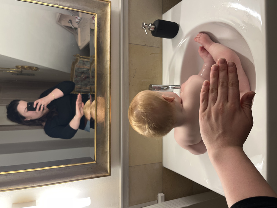 The scene depicts a bathroom interior located in Hohenbrunn, Germany. A fair-skinned infant with light reddish-blonde hair is positioned in a white rectangular sink, facing away from the viewer towards a chrome-finished faucet. Water is visible in the sink basin around the infant's submerged lower body. An adult's bare forearm and open hand are visible in the immediate foreground, positioned over the infant's back. To the right of the sink, a black, pump-style soap dispenser with a chrome top is present. Mounted above the sink is a large mirror with an ornate, gold-toned frame. The mirror reflects an adult female, identified as the photographer, holding a smartphone. She has dark hair and is wearing a dark, long-sleeved top. The reflection also reveals a wall-mounted light sconce on the left and partially visible furniture, possibly a dresser or cabinet, in the distant background.