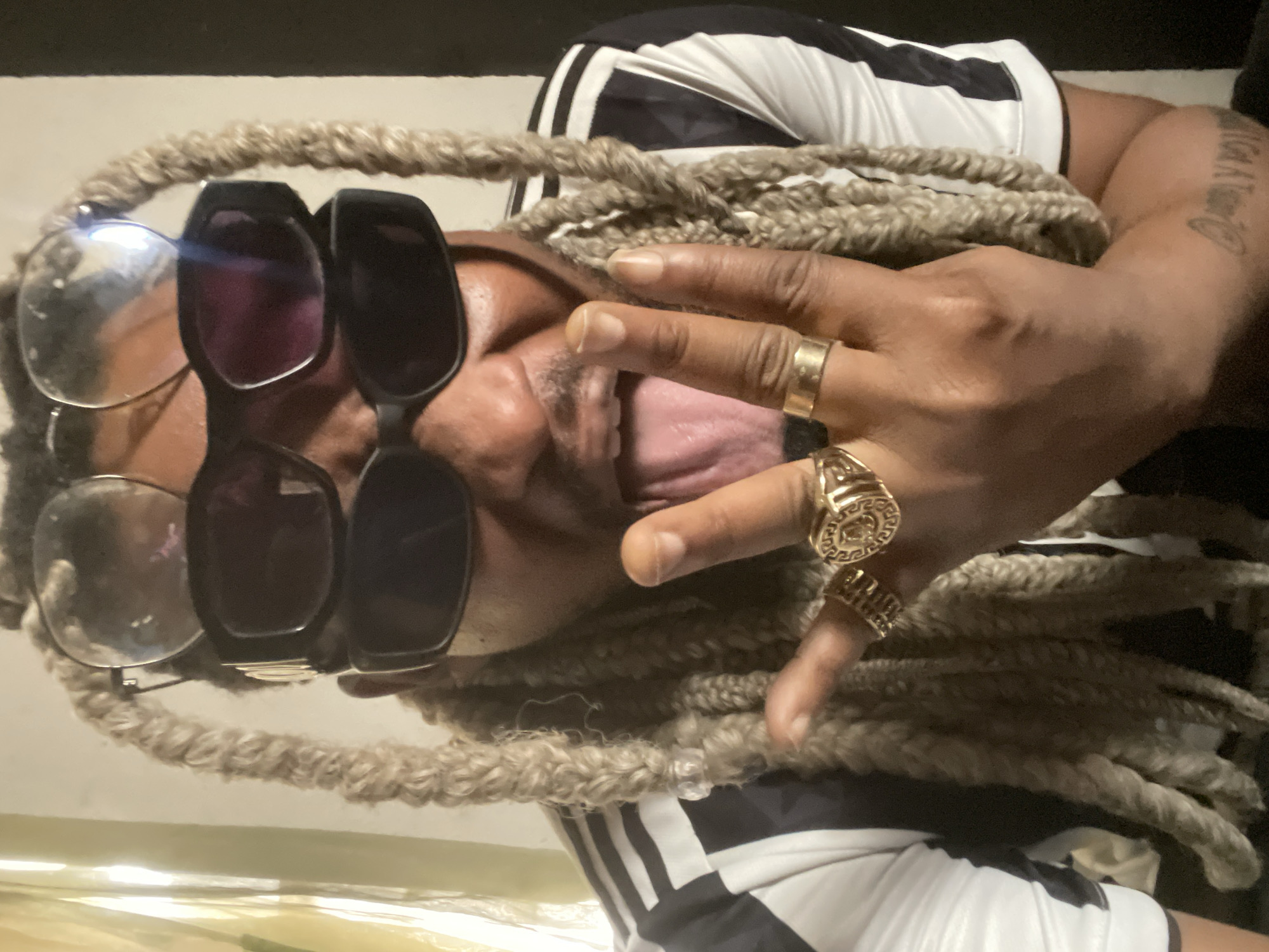 A person with light brown dreadlocks, wearing a black and white striped shirt, poses with two pairs of glasses (one clear, one sunglasses) and several gold rings. The person is sticking their tongue out and facing sideways. The image was taken indoors. The city of Jos, Nigeria is not discernible from the image.
