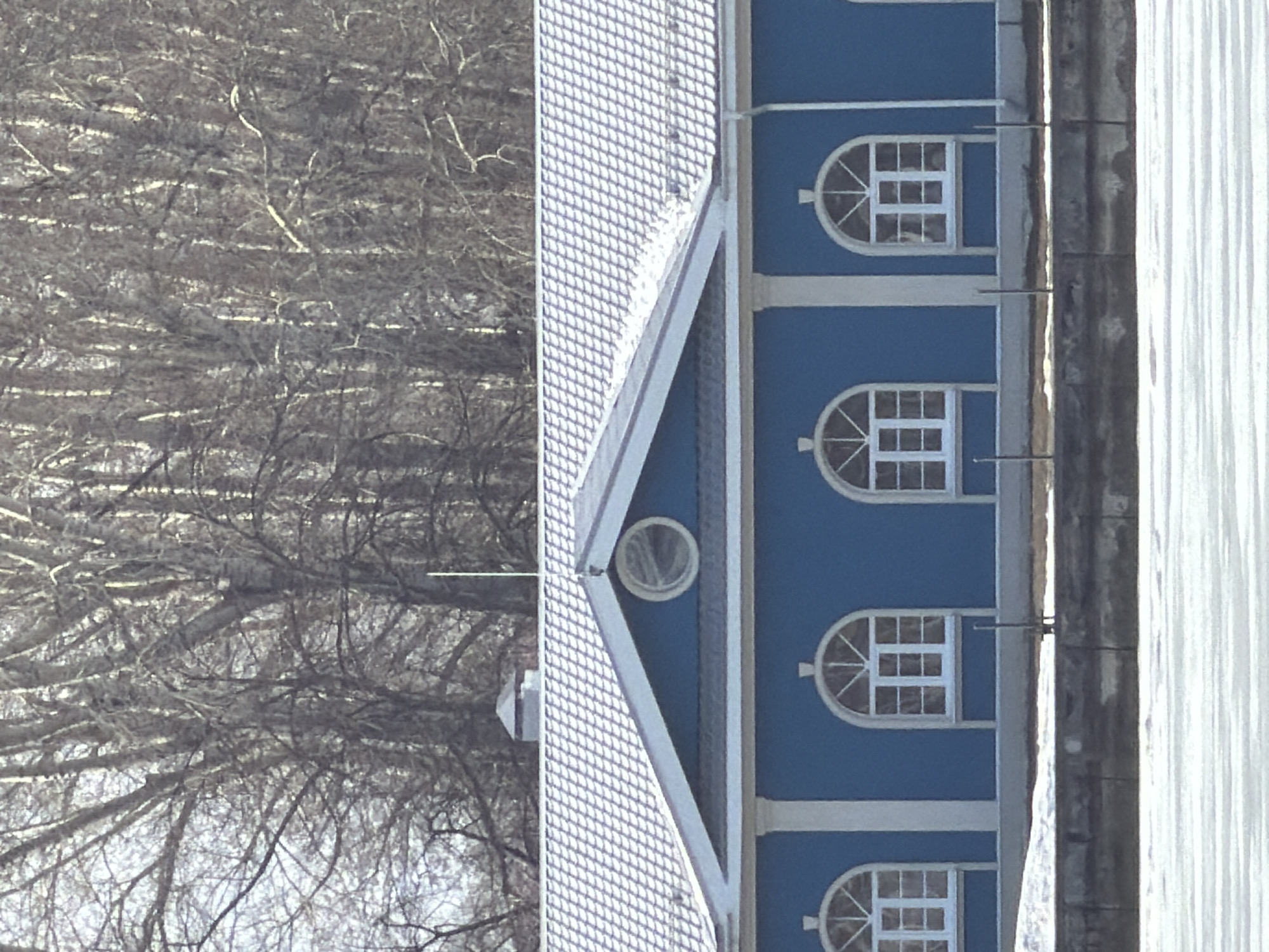 The image shows a building in Votkinsk, Russia. The building has a blue facade with white trim and a grey roof. There are several arched windows on the facade. A forest is visible in the background. The ground is covered in snow.