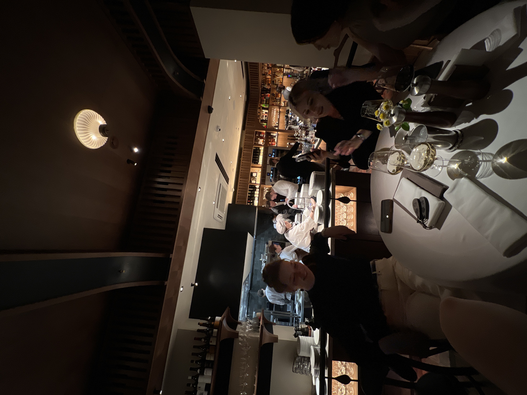 This sideways-oriented image captures the interior of a stylish, dimly lit restaurant or bar, likely situated in Moscow, Russia, during the evening. The setting exudes an upscale and intimate ambiance with its warm lighting and modern design.
In the immediate foreground, a diner with curly hair is smiling at the camera, wearing a dark top. To their right, at a round, white table, two other diners are visible. The table is elegantly set with multiple wine and water glasses, a small vase holding yellow flowers, and metallic salt and pepper shakers. One person is engrossed with their phone.
Further into the background, an open kitchen concept is prominent, where several chefs in white uniforms are actively working, preparing food. Adjacent to this culinary area is a well-stocked bar featuring numerous bottles of various spirits and wines displayed on wooden shelving. Another staff member is seen behind the bar.
Architectural details include dark wooden paneling on the walls and ceiling, creating a sophisticated backdrop. A distinctive circular ceiling light fixture with a ribbed design emits a soft, warm glow, enhancing the overall inviting atmosphere. The scene depicts a lively yet refined dining experience. No visible text is clearly legible within the image.