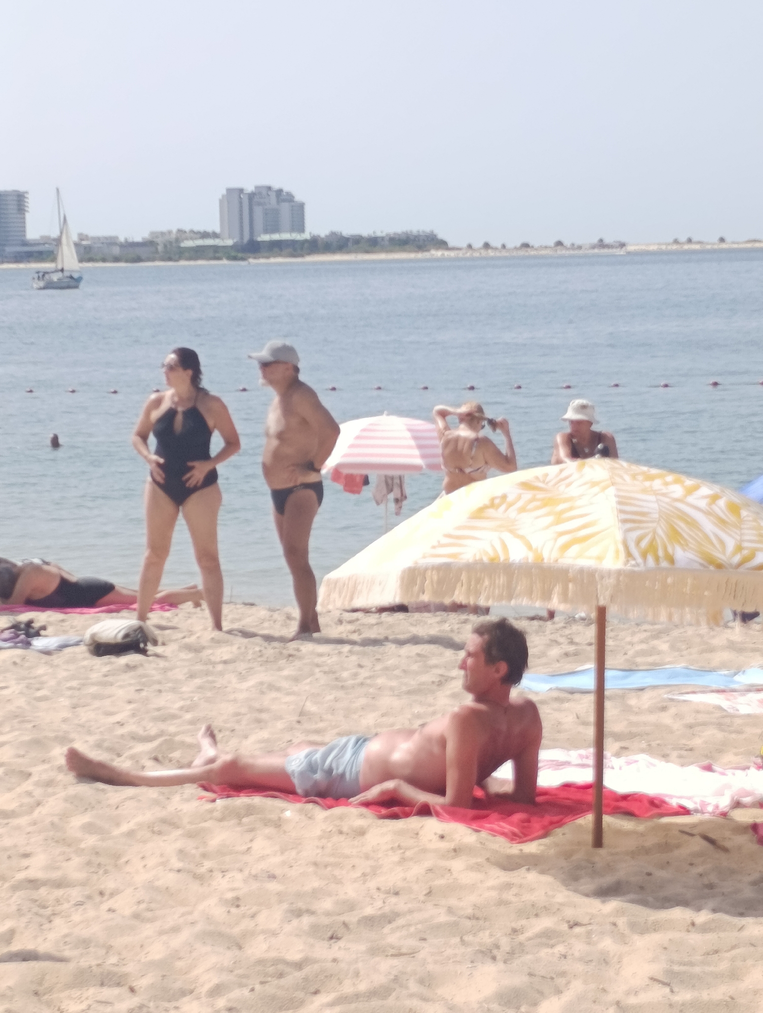 This is a photograph taken from a beach in Setúbal, Portugal, with the city's coastline visible in the background. In the foreground, a man is lying on a red towel on the sand, facing to his left. He is wearing light blue swim trunks. Behind him, a yellow and white striped umbrella is set up. Further back, two people are standing near the water's edge. The woman on the left is wearing a black one-piece swimsuit and sunglasses, with her hands on her stomach. The man next to her is wearing a light-colored cap and dark swim trunks. In the middle ground, a sailboat is visible on the water. The background shows a hazy cityscape with buildings and a clear sky. The overall scene depicts a sunny day at the beach with several people enjoying the environment.