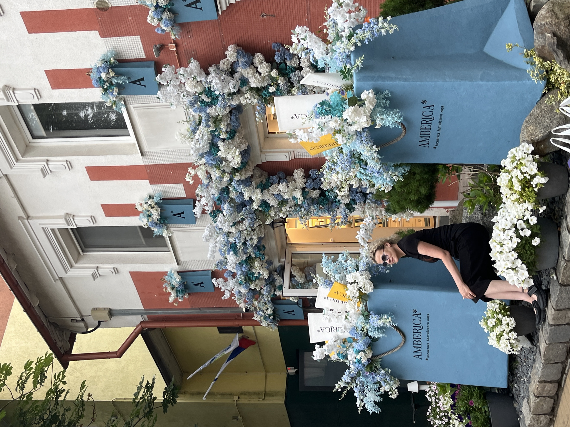 A woman with blonde curly hair, wearing sunglasses and a black outfit, sits on a stone step in front of a storefront in Zelenogradsk, Russia. The storefront is decorated with an abundance of blue and white artificial flowers cascading over an arched entryway. Two large light blue, bag-shaped displays with the text "AMBERICA* Косметика Балтийского моря" (AMBERICA* Baltic Sea Cosmetics) are placed to the left and right of the entrance. The woman is looking to her right. In front of the displays are potted white flowers. The building facade is red with white trim and two windows. Russian flags are visible on an adjacent yellow building. The overall scene suggests a retail establishment focused on cosmetics, possibly leveraging the Baltic Sea location for branding.