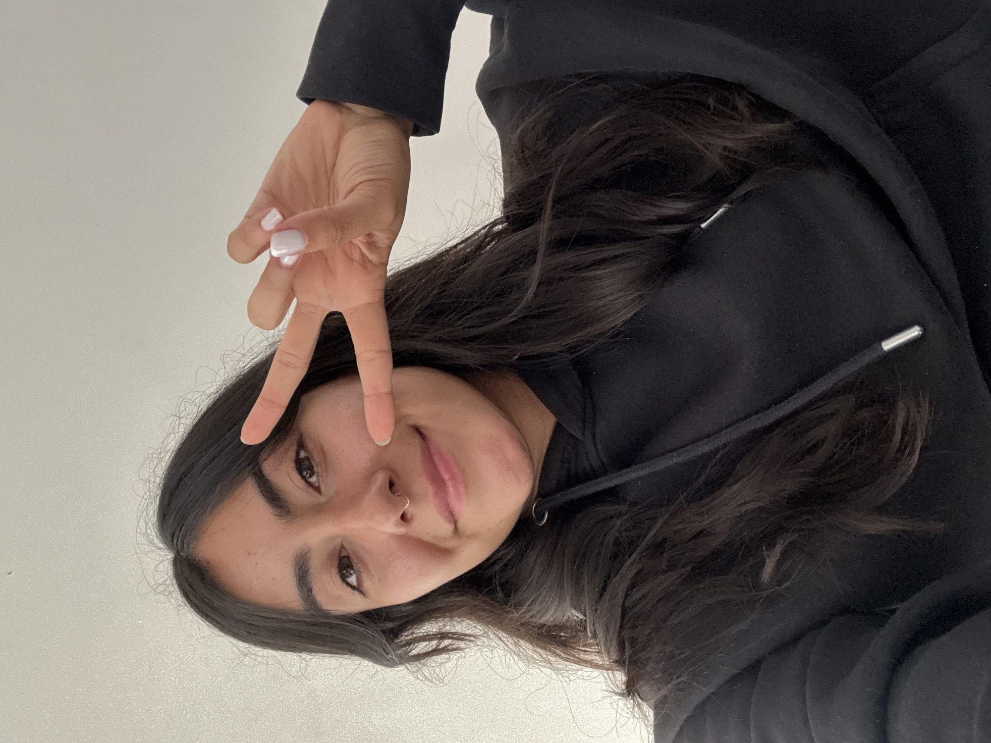 A young woman wearing a black hooded sweatshirt poses for a selfie with her hand held up in a peace sign. She has dark hair and a nose ring. The background is a plain white wall. The location is unknown and cannot be determined from the image.