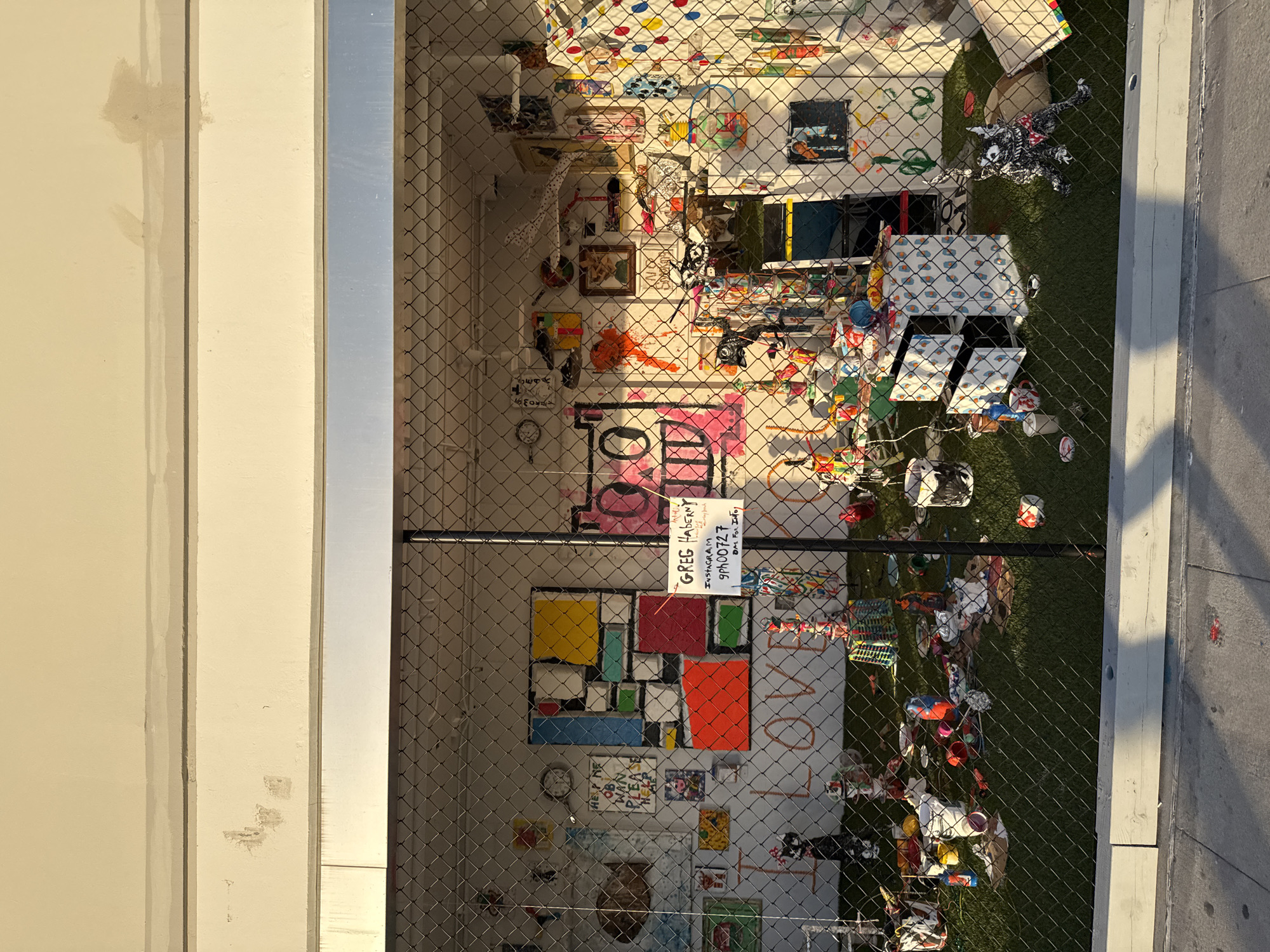 The image shows an enclosed space, possibly an outdoor room or display area, behind a chain-link fence. The location is likely Los Angeles, California, as indicated by the style of architecture and vegetation.
Inside the fenced area, there is an eclectic and chaotic collection of items. The back wall is adorned with colorful artwork, including a Mondrian-inspired composition of squares and rectangles. Other pieces of art and various objects are scattered throughout the space.
A sign attached to the fence provides the name "Greg Haberny" along with an Instagram handle ("9PN00727"). The phrase "DM For Info" is also on the sign.
The floor of the enclosed space appears to be covered with artificial turf, with various objects and debris scattered across it. These objects include what looks like toys, containers, and other miscellaneous items.
The overall impression is one of artistic expression and playful disarray.
