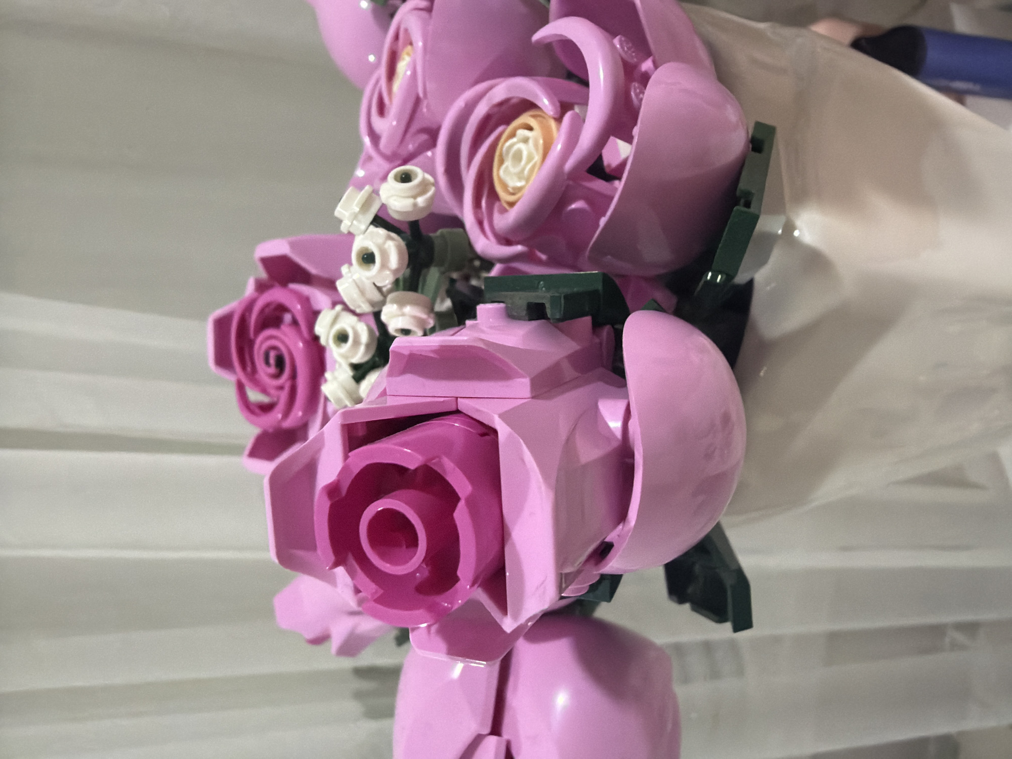 A close-up photograph captures a bouquet of constructed pink and white floral objects, identified as LEGO-type bricks, presented in a white vase, located in the city of Dobrich, Bulgaria. The primary subjects are several large, stylized pink roses, with varying degrees of openness. Some roses display a central spiral pattern in a deeper magenta, while others feature a light beige or white inner core. Interspersed among the pink roses are smaller clusters of white floral objects with dark centers, resembling filler flowers such as baby's breath. Dark green LEGO elements are visible, forming the structural stems and leaves supporting the bouquet. The flowers are arranged within a white, matte-finish vase, of which the upper portion is visible. The background consists of a uniformly light-colored, vertically pleated fabric, likely curtains or drapes, rendered out of focus. No individuals, overt actions, or interactions are present in the frame.