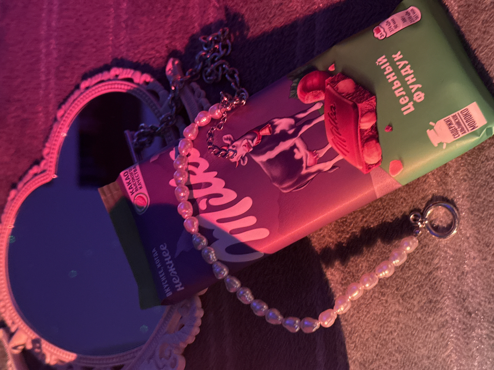 The image presents a still life composition, dominated by a Milka chocolate bar package resting on a soft, textured surface. The package is purple on one side, adorned with the Milka cow logo, and green on the other, labeled "Цельный Фундук". A small, heart-shaped mirror with a decorative frame is situated to the left, reflecting a deep blue hue. A pearl necklace, featuring irregular pearl shapes, is draped across the chocolate bar, with a silver chain necklace intertwined nearby. The lighting in the image is predominantly red and blue, creating a dramatic, contrasting effect and casting strong shadows. The OCR identifies Russian text related to the chocolate bar's attributes, emphasizing its superior quality and content.