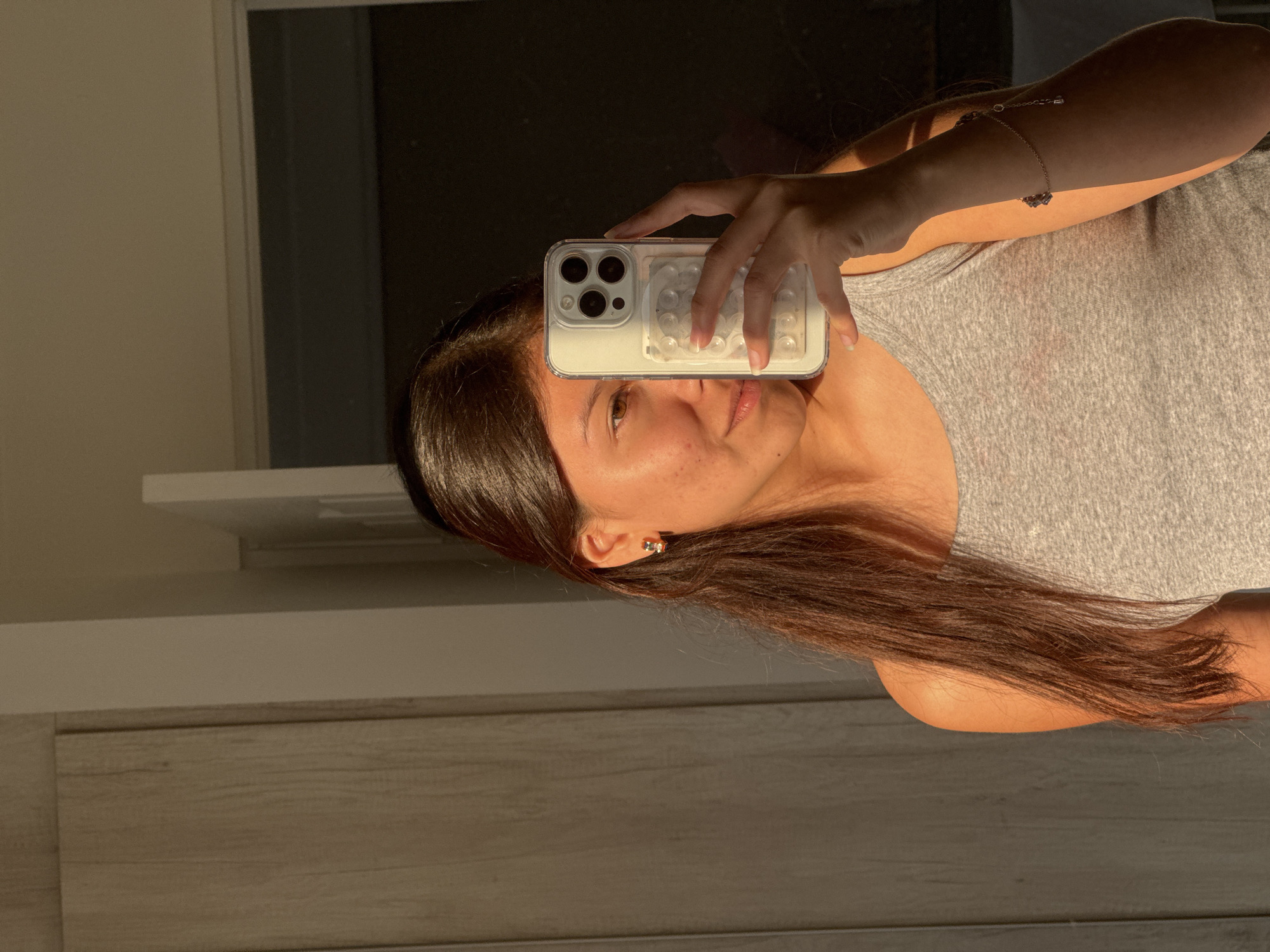 A woman with long, dark brown hair is holding a white iPhone with a case featuring a pattern of clear, raised circles in front of her face, partially obscuring it. She is wearing a gray tank top and a delicate bracelet on her left wrist. The background includes a partially visible window and what appears to be a light-colored wooden cabinet or wall. The lighting suggests an indoor setting with sunlight streaming in, casting shadows. There is no verifiable evidence to confirm that the location is in the city of San Francisco Ocotlán (Ocotlán), Mexico.