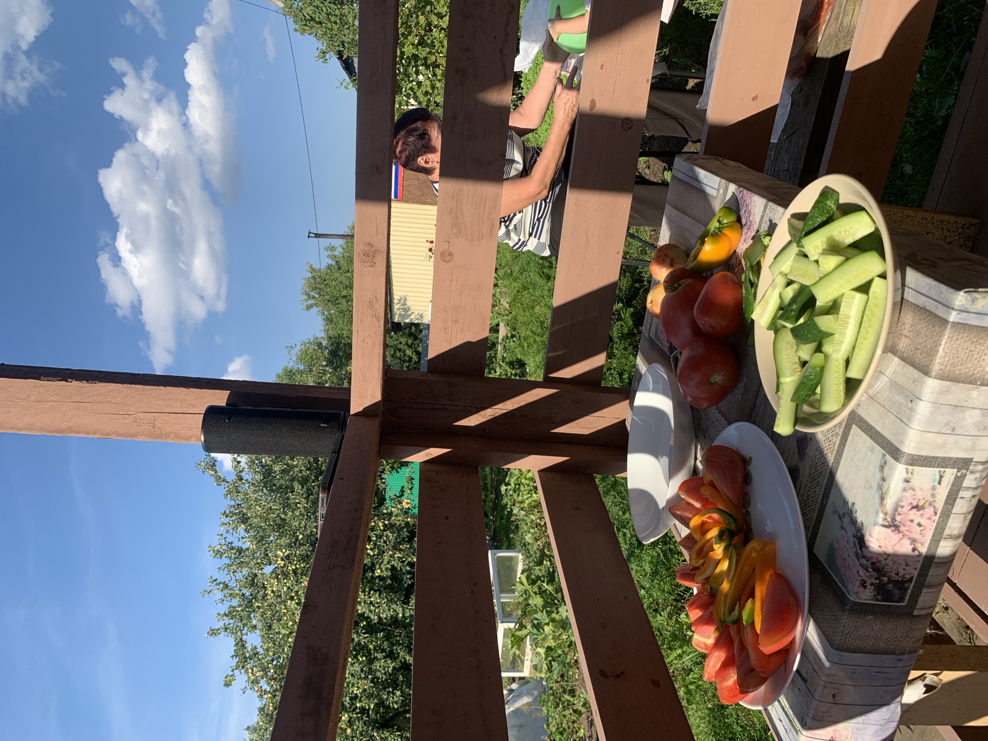 The image depicts a rustic outdoor scene, likely a backyard or dacha area, under a clear blue sky with scattered white clouds. In the foreground, a table is set with a colorful tablecloth. On the table, there are two plates of fresh vegetables: one with sliced tomatoes and peppers, and another with sliced cucumbers. Several whole tomatoes and apples are also visible on the table. A white bowl rests behind the plate of tomatoes.
A woman with short, dark hair and wearing a striped t-shirt is partially visible in the background, appearing to be engaged in some activity. A black cylindrical speaker is placed on a wooden beam in the upper left portion of the image, atop a smartphone. The background reveals a garden with lush green foliage and trees, suggesting a rural or suburban setting. The overall impression is that of a casual outdoor meal preparation or gathering on a sunny day. The location is identified as Chaikovsky, Russia.