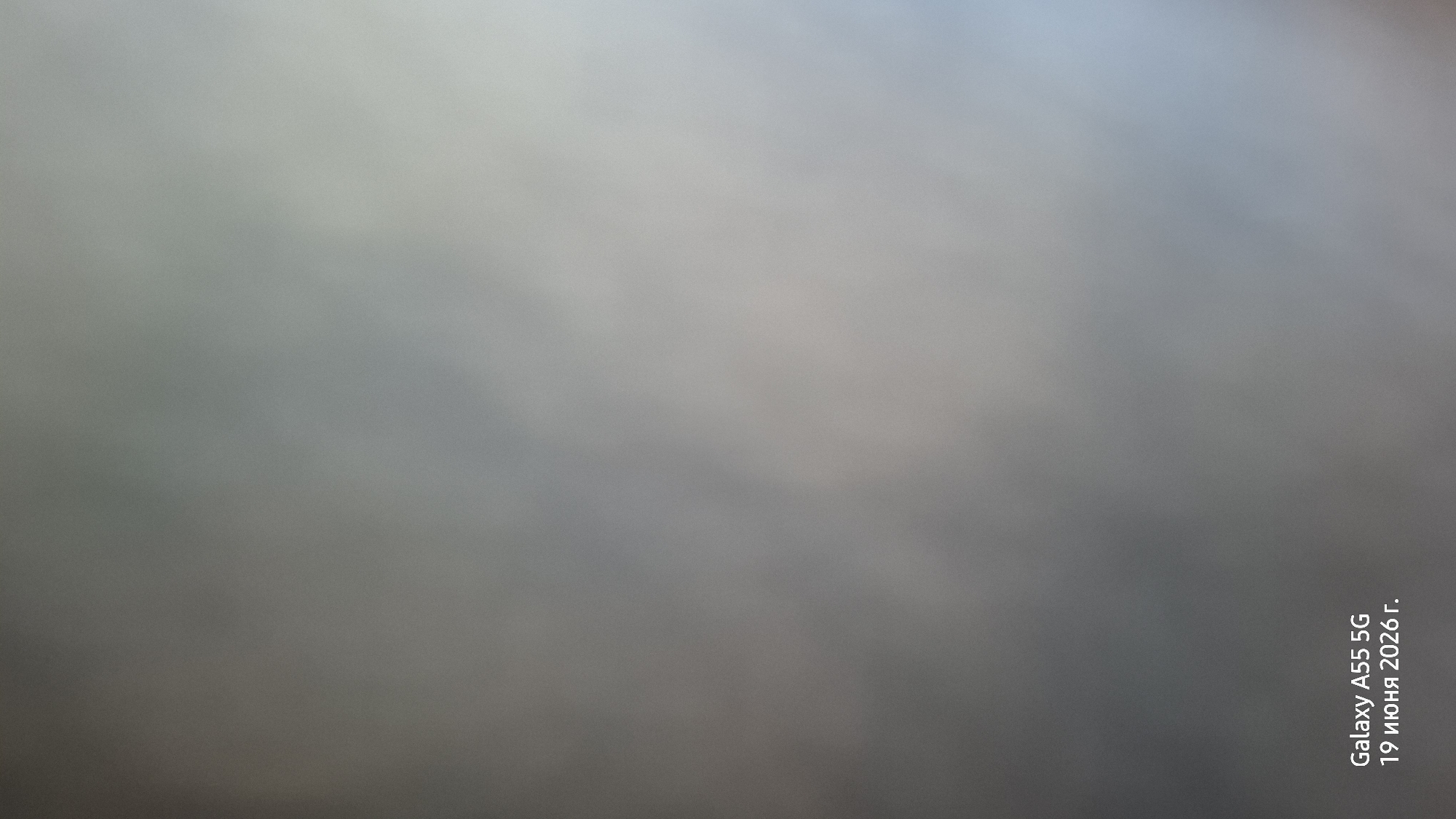 The image is an abstract, out-of-focus shot with a light grey and blue gradient. The overall impression is soft and hazy. In the bottom right corner, oriented vertically, there is white text that reads "Galaxy A55 5G 19 июня 2026 г.". This text indicates the device used to capture the image and the date, June 19, 2026. Due to the lack of distinct features, it's impossible to determine the specific subjects, activity, time of day, weather, or location beyond what the text provides.