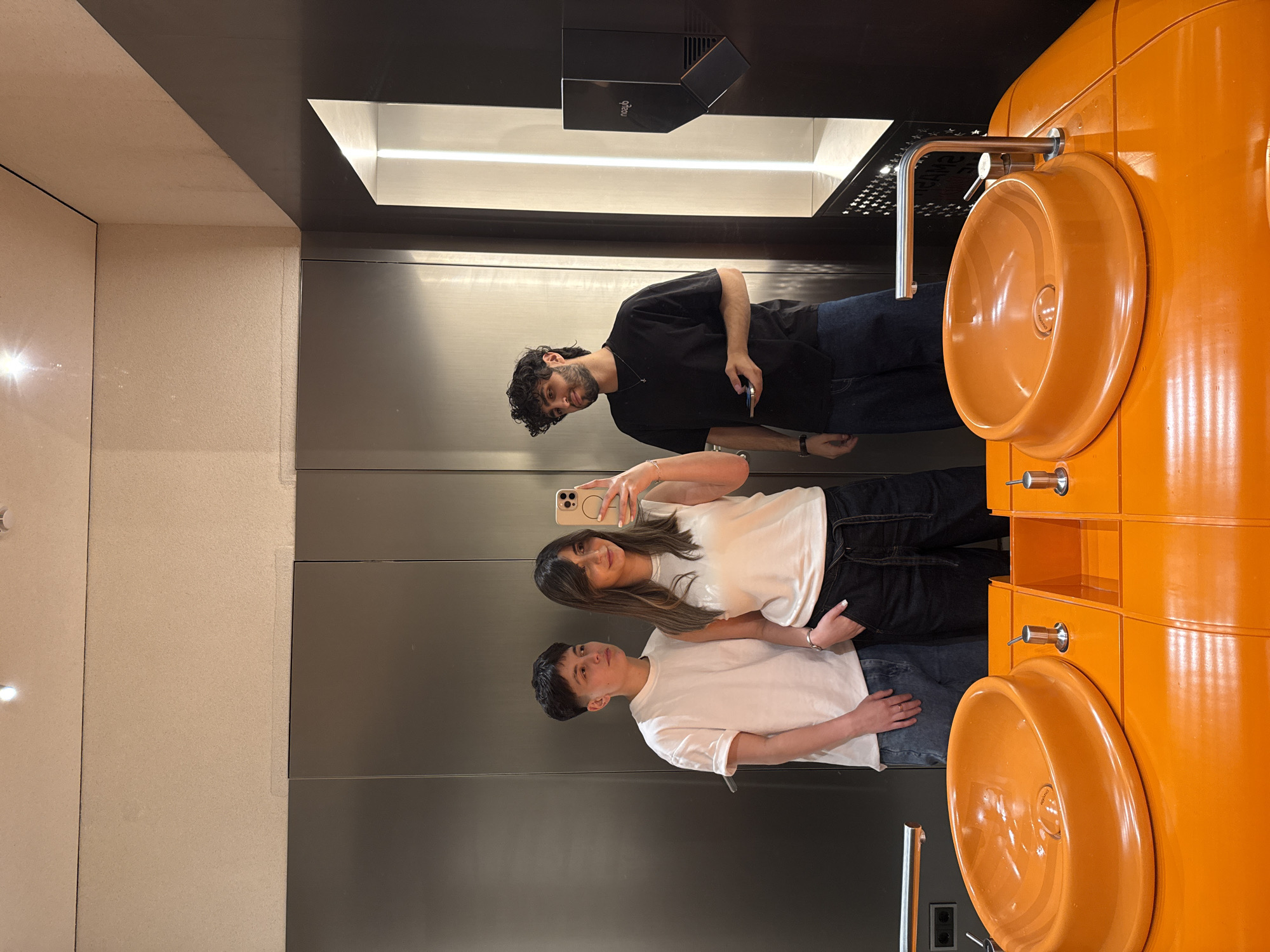 Three individuals, two males and one female, are captured in what appears to be a mirror selfie taken within a modern washroom. The image is oriented sideways, but if viewed upright, the subjects are standing side-by-side. The female in the center, wearing a white t-shirt and dark pants, holds a smartphone to take the picture. To her left stands a male with dark curly hair and a beard, wearing a black t-shirt and jeans, also holding a phone. To her right, a second male in a white t-shirt and lighter jeans is visible.
The setting is a contemporary washroom characterized by a distinctive, bright orange vanity unit featuring two circular sinks with chrome faucets, positioned to the right of the individuals. The walls behind them are a combination of sleek, reflective dark panels and lighter, textured surfaces. Above the reflective surface, a horizontal light strip provides illumination. A black rectangular device, possibly a diffuser or air quality sensor, is mounted on a shelf above the lighting, displaying reversed text that appears to be "qbcu." Faint reversed text, "mondo," is also visible in the basin of one of the sinks. The casual attire of the individuals and the striking design of the washroom suggest a public or commercial establishment in Yerevan, Armenia.