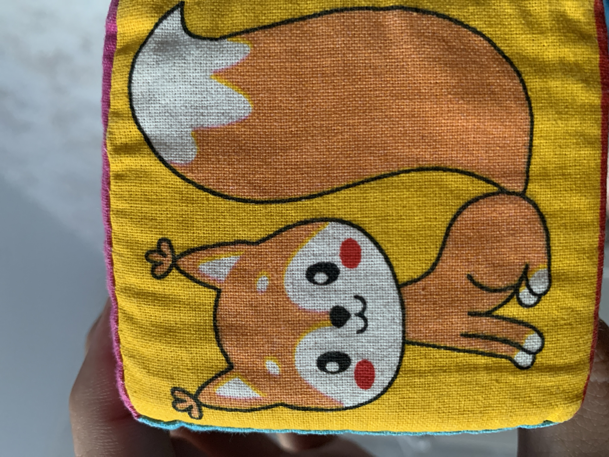 The image shows a close-up of a yellow fabric cube featuring a cartoon drawing of a fox. The fox is depicted lying on its back with its tail curled over its body. The tail is orange with white markings and a black outline. The fox's body is mostly orange and white with black outlines. It has a white face with black eyes, a small black nose and mouth, and two red circles for cheeks. Its paws are also outlined in black with white tips. The fabric cube has pink and blue trim along its edges. The overall impression is of a children's toy or decorative item. The setting appears to be indoors, with a neutral background that suggests a surface like a table or floor. There is no visible text in the image. The image is well-lit, suggesting it was taken in daylight or under artificial lighting. The orientation of the image is slightly tilted.