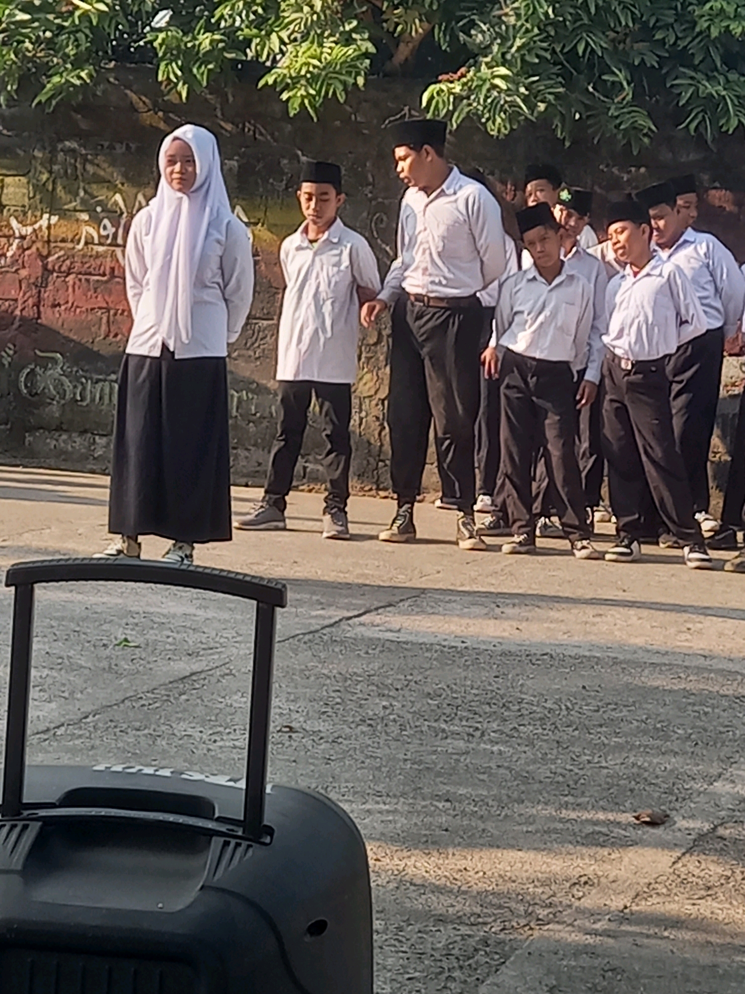 The image depicts a group of children, some wearing religious headwear, standing in what appears to be an outdoor educational setting, possibly in Perampuan, Indonesia. The children are dressed in white shirts and dark pants or skirts. One child in the foreground wears a white hijab and a dark skirt. Behind them, a brick wall with some markings is visible, and lush green foliage hangs overhead. The ground is paved with concrete. In the foreground, the top portion of a black rolling object, likely a speaker or equipment case, is present, suggesting an event or gathering. The lighting indicates it is daytime, possibly morning or late afternoon, with sunlight casting shadows.
